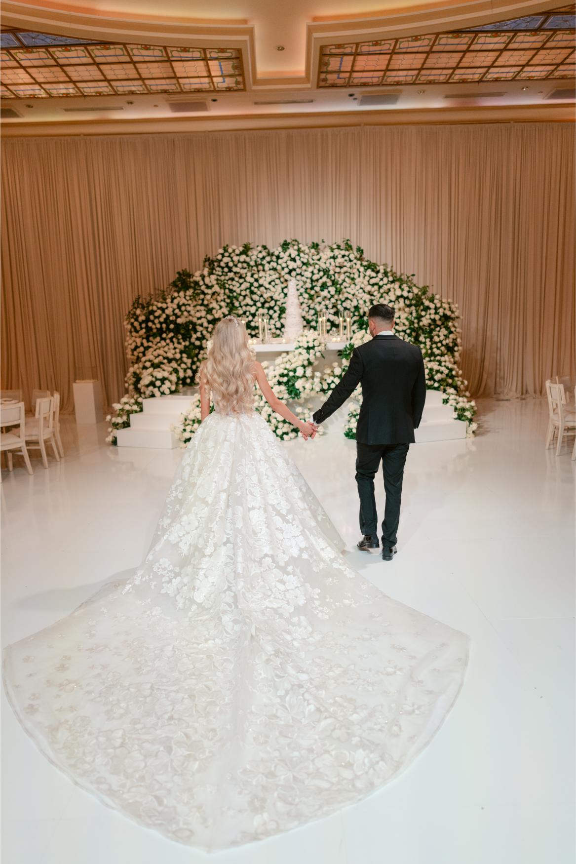 Couple holding hands in a wedding ceremony with a floral arch in the background. Bride Anaies wearing the Renee from Galia Lahav's Rise Collection