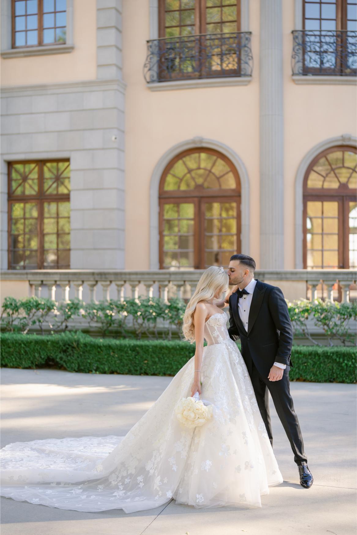 Couple in wedding attire standing in front of a large building with arched windows. The Bride, Anaies, wearing the Renee from Galia Lahav's Rise Collection