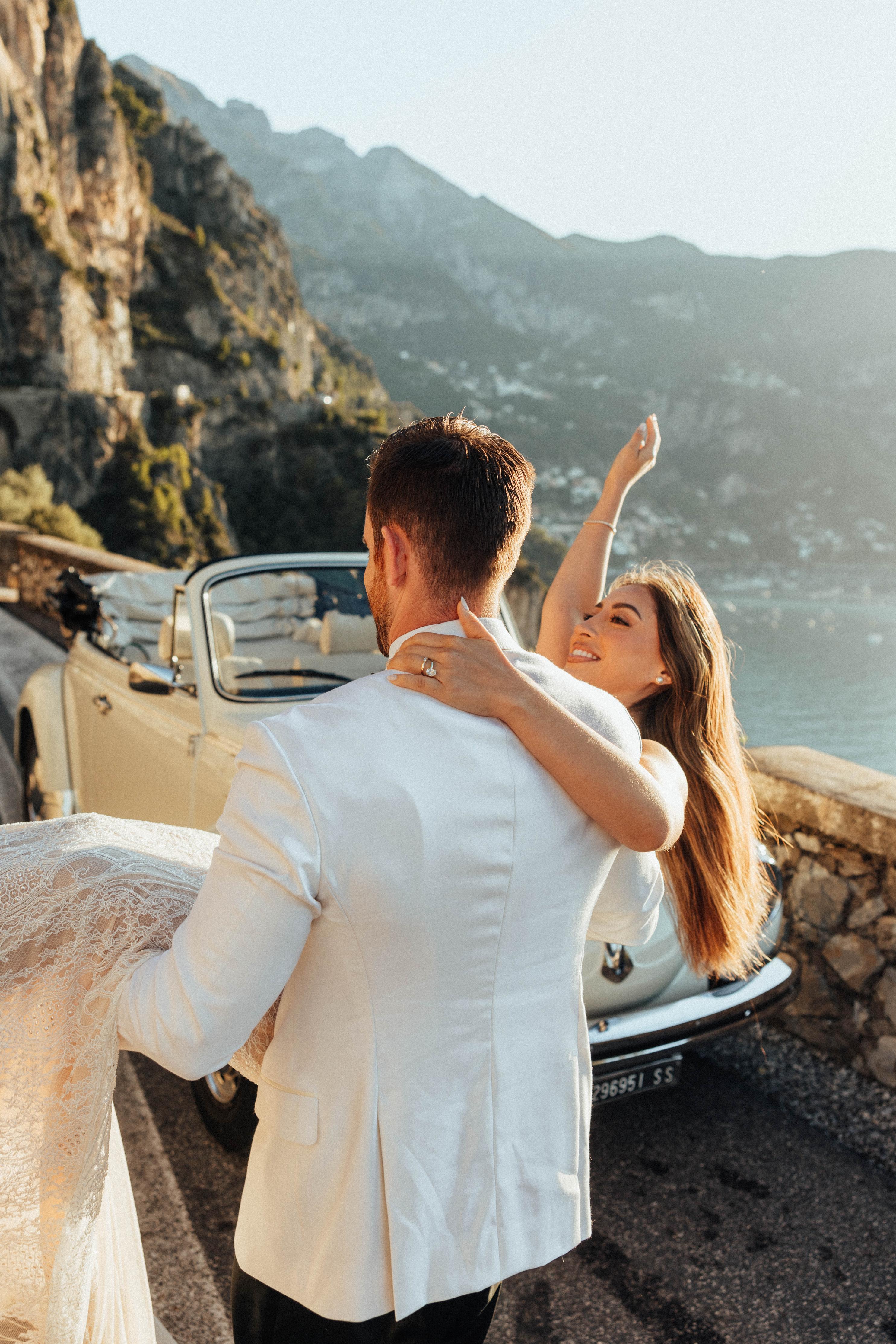 GL Bride Betty Luna in our Élysée being carried by her husband with a vintage car and seaside in the background