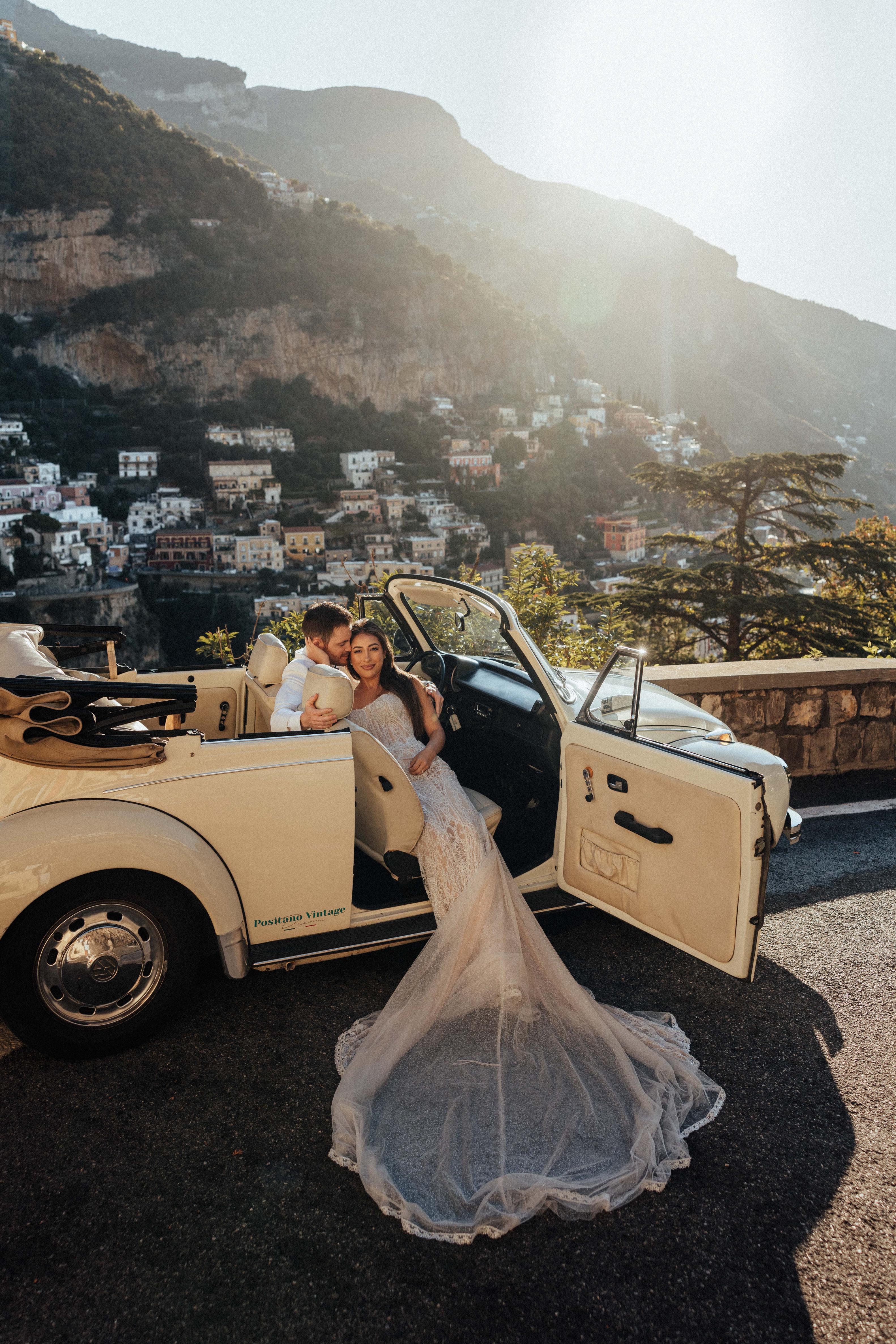 GL Bride Betty Luna in our Élysée with her husband sitting in a vintage car, by a cliffside city overlooking the sea 