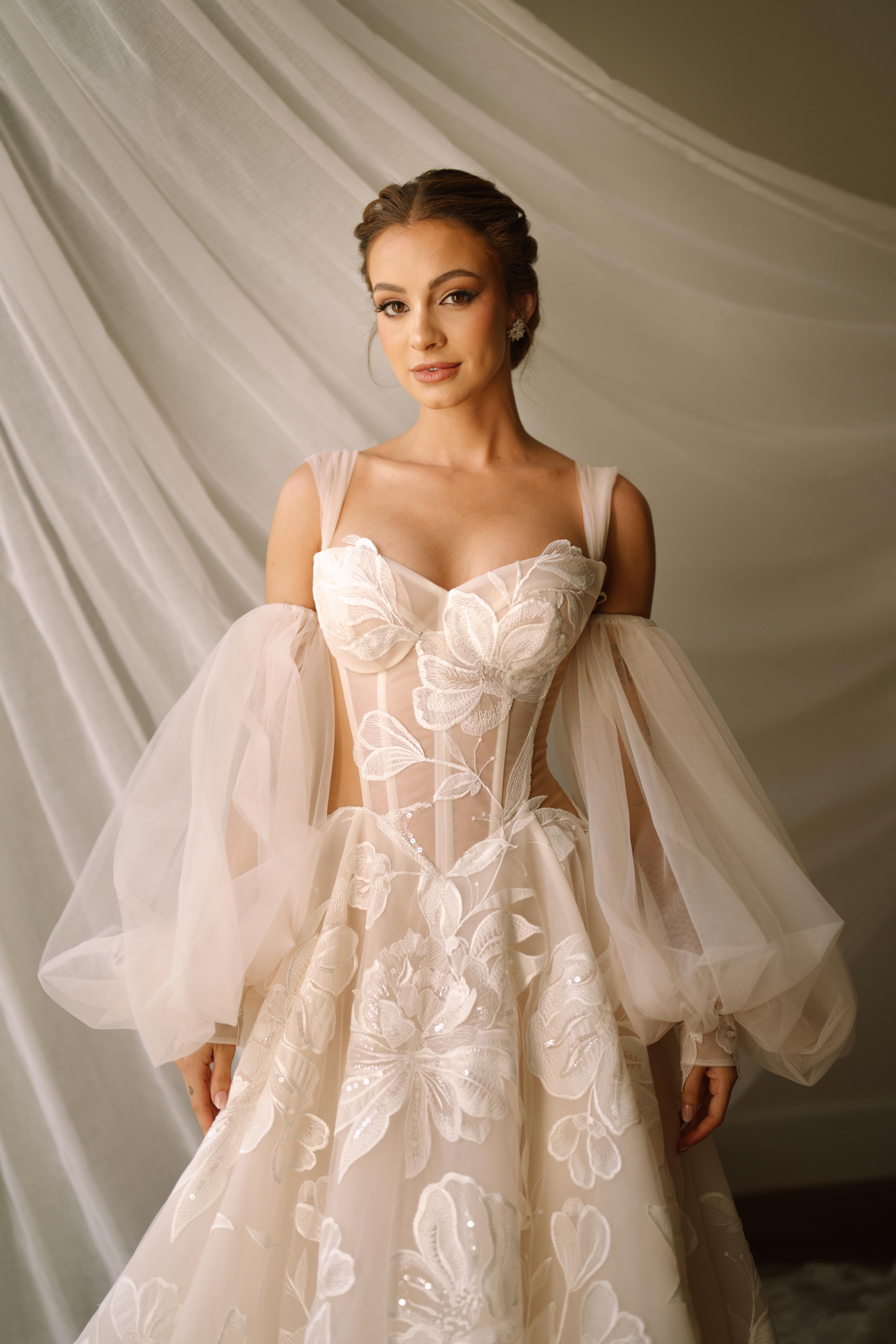 GL Bride Carina wearing the Evelyn wedding dress with floral details against a soft, draped fabric background.