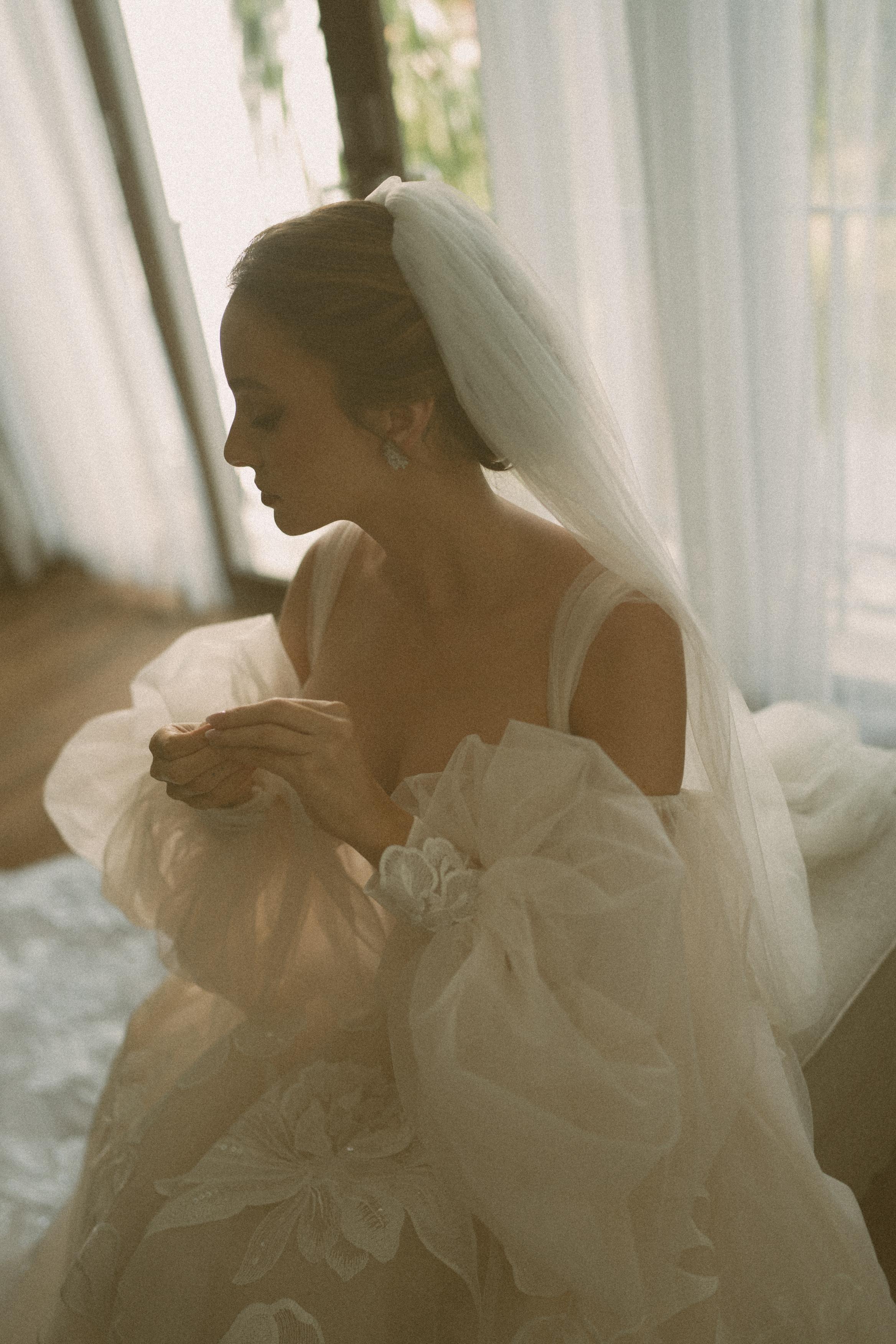 GL Bride Carina wearing the Evelyn wedding gown with a veil, sitting by a window.