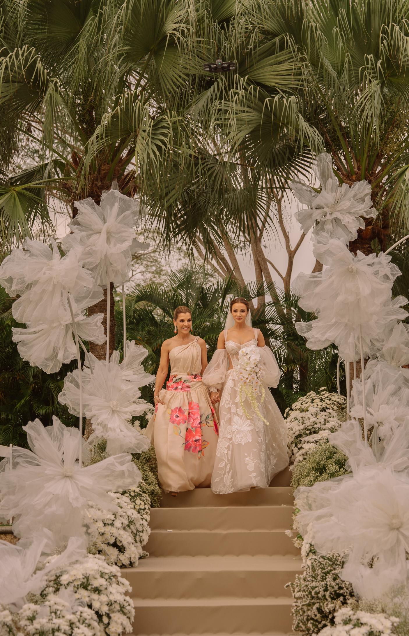 Two women in elegant dresses standing on a decorated pathway with large white flowers and greenery.