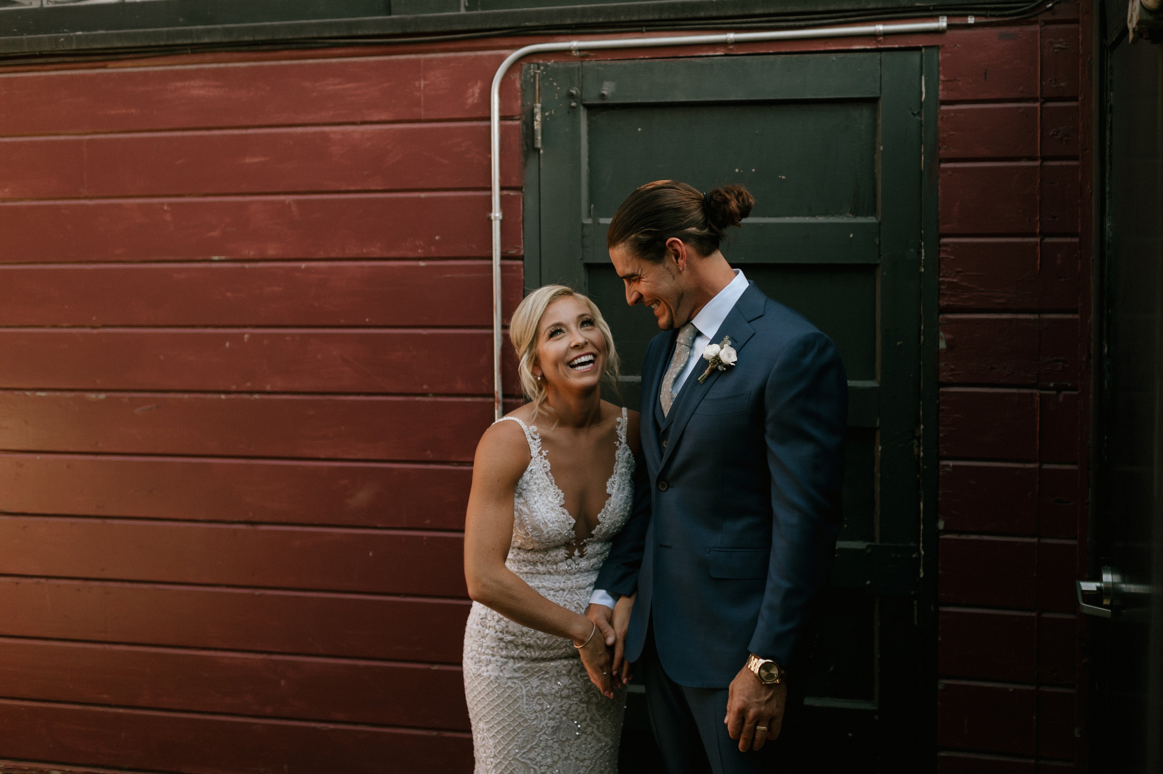 GL Bride Carly wearing our GALA 708 with her husband, laughing in front of a wooden building 