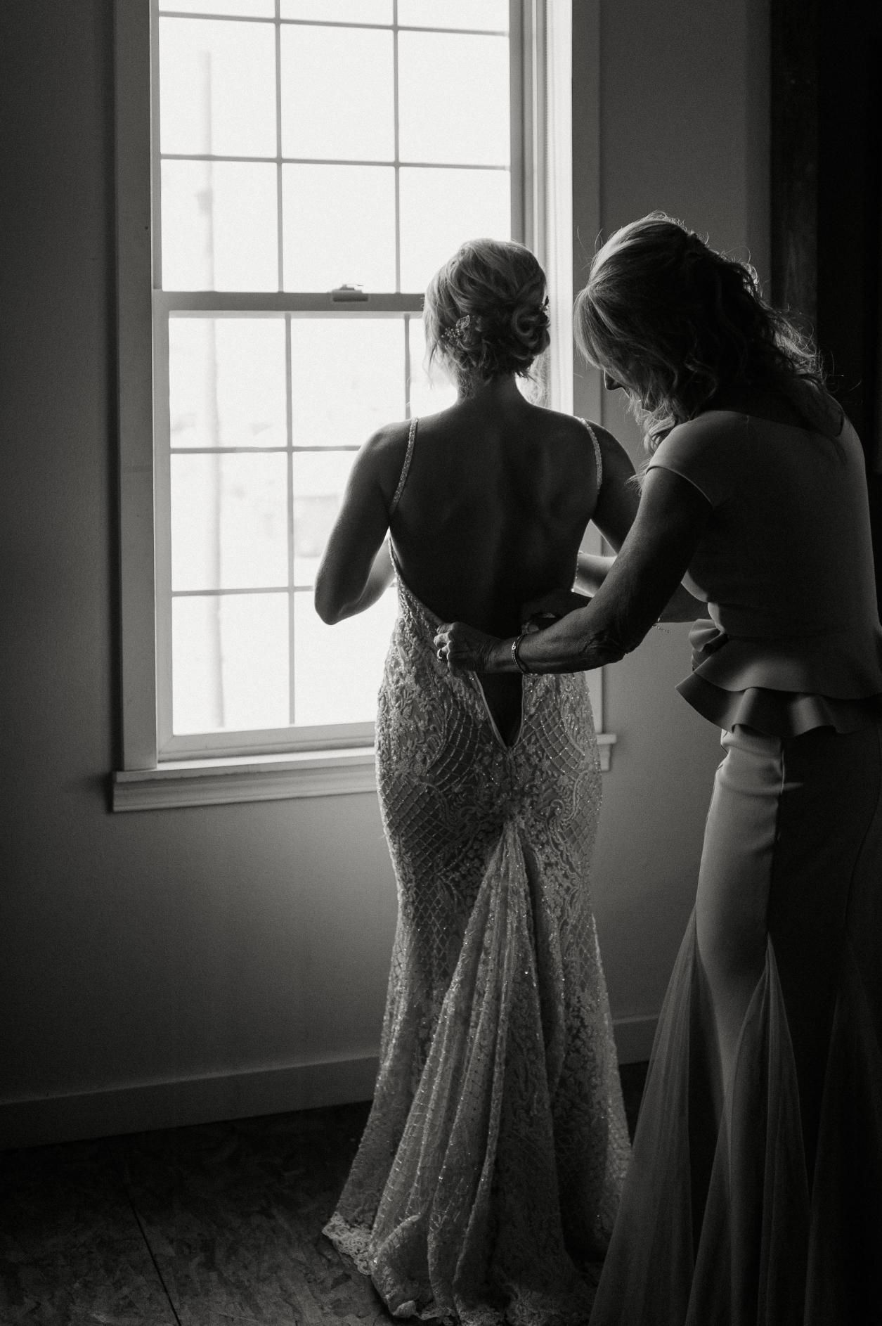 Black and white photo of a woman helping our GL Bride Carly into the GALA 708 dress by a window.