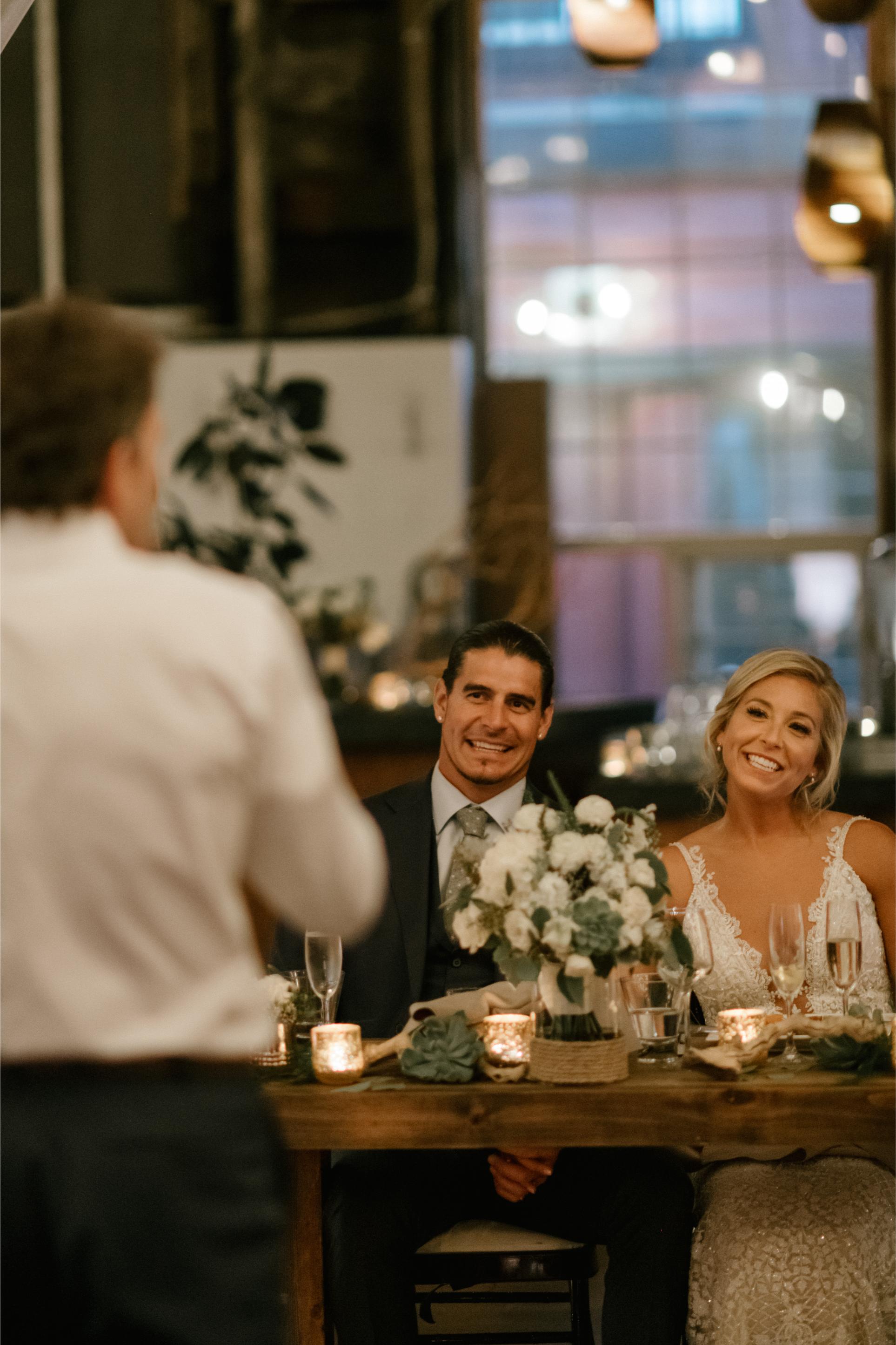 GL Bride Carly in the GALA 708 sitting with her husband at a table with floral arrangements and candles, smiling towards the camera.