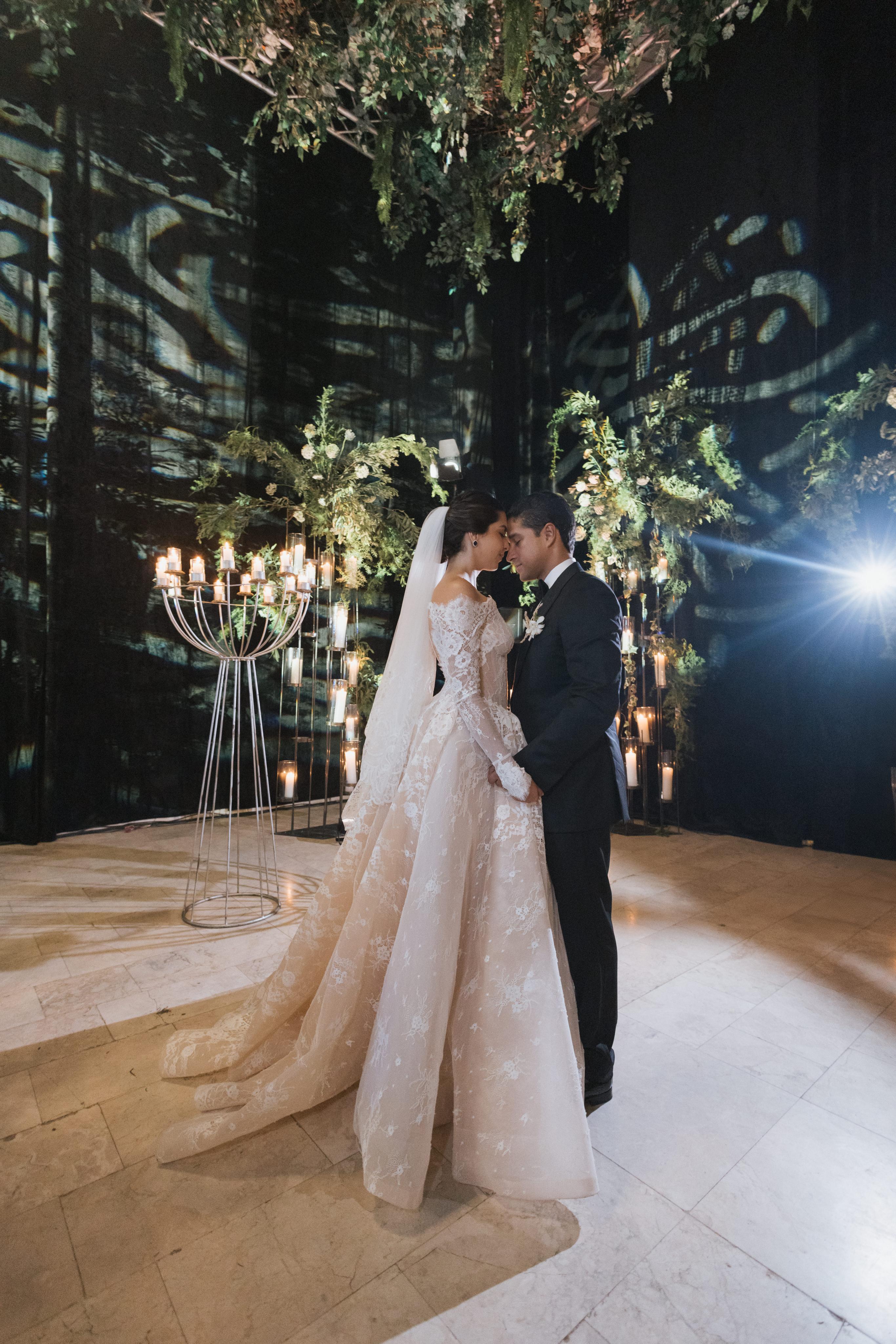 Couple in wedding attire standing in a decorated indoor setting with greenery and lights.