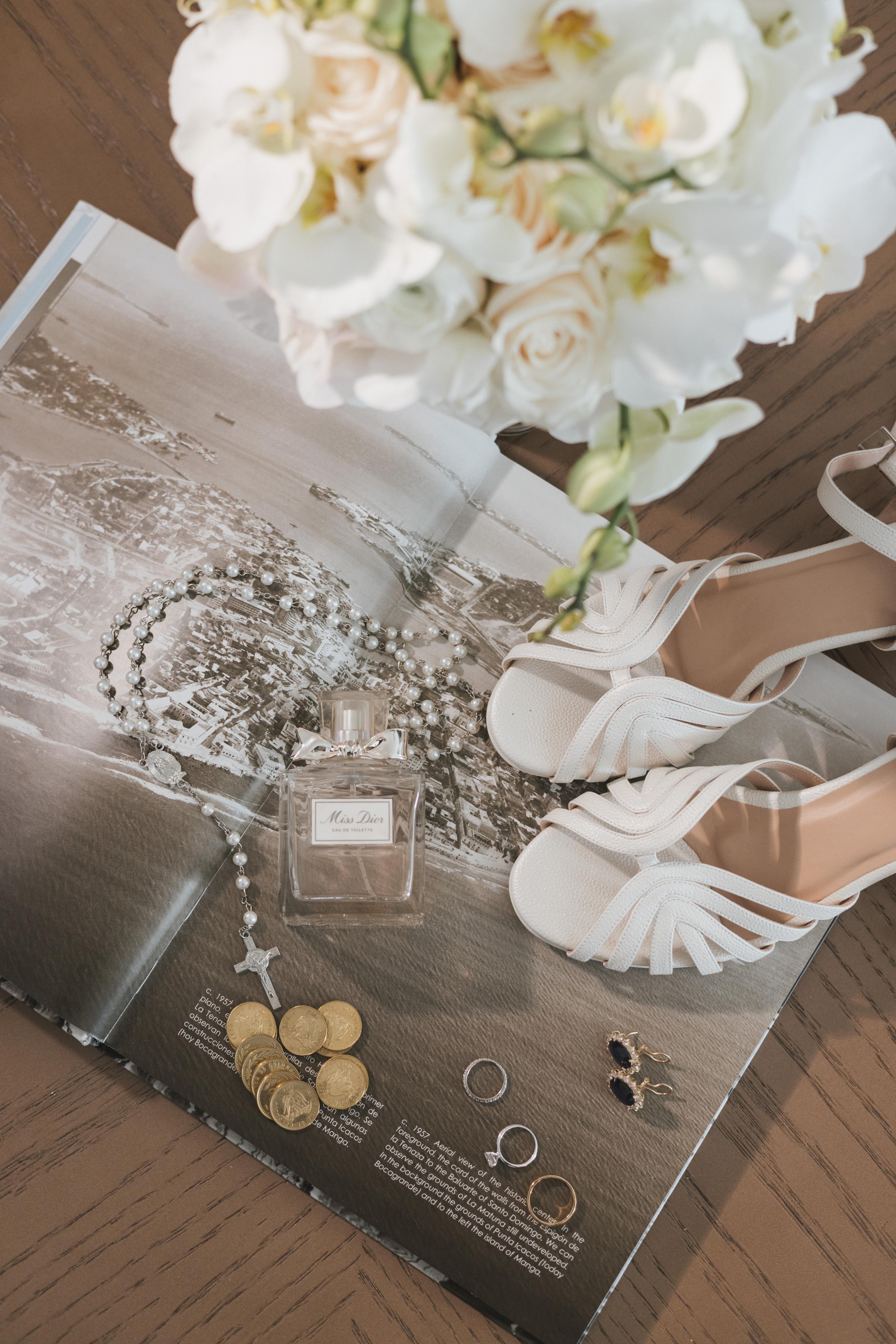 White wedding shoes next to a magazine with a perfume bottle and flowers on a wooden surface