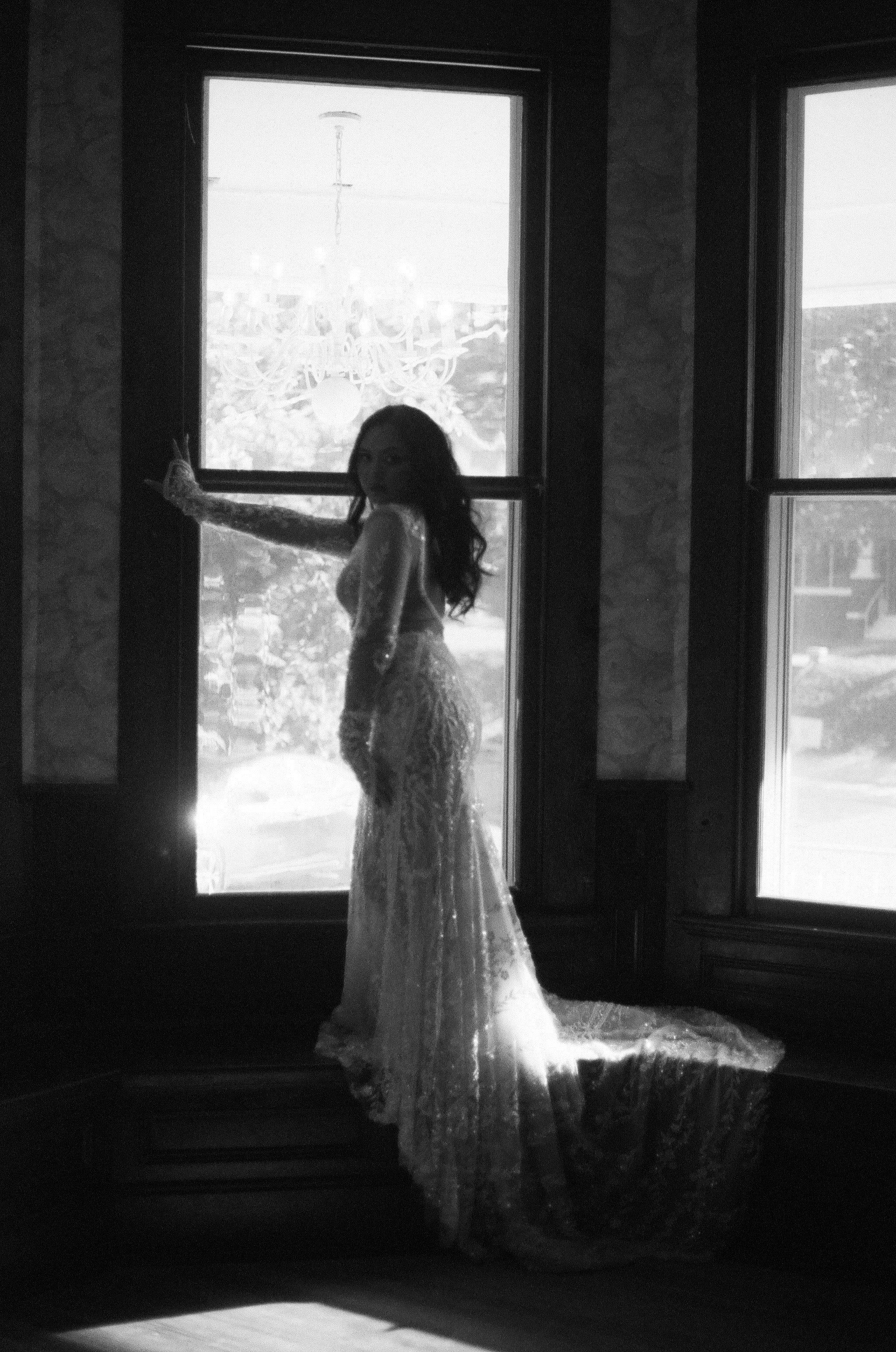 Woman in a flowing dress standing by large windows with sunlight streaming through.