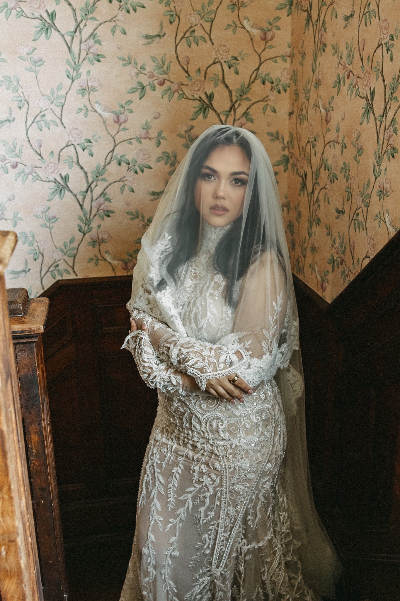 Woman in a lace wedding dress with a veil standing against a decorative wall.