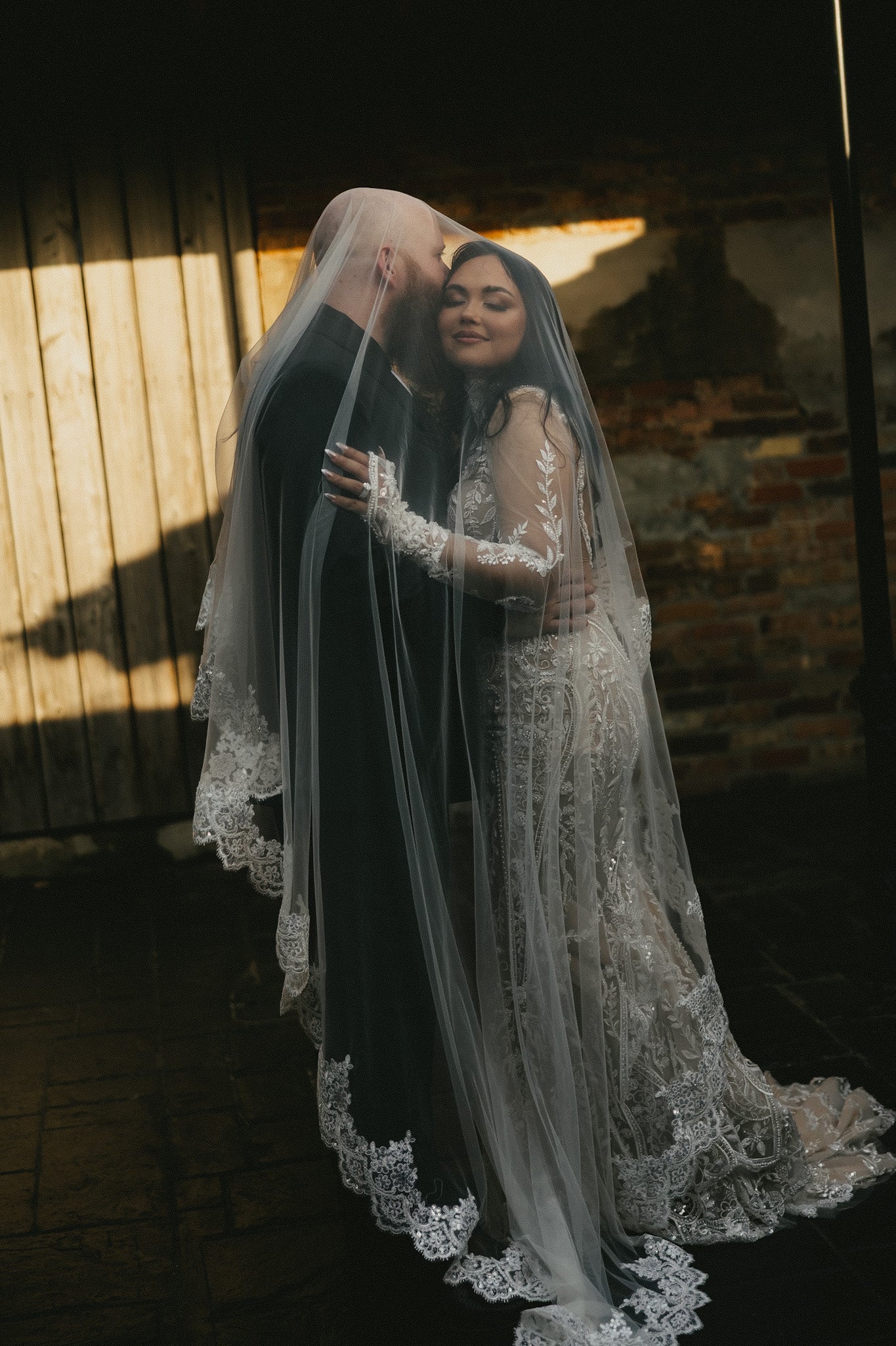 Two people embracing with a long veil in front of a rustic wall.