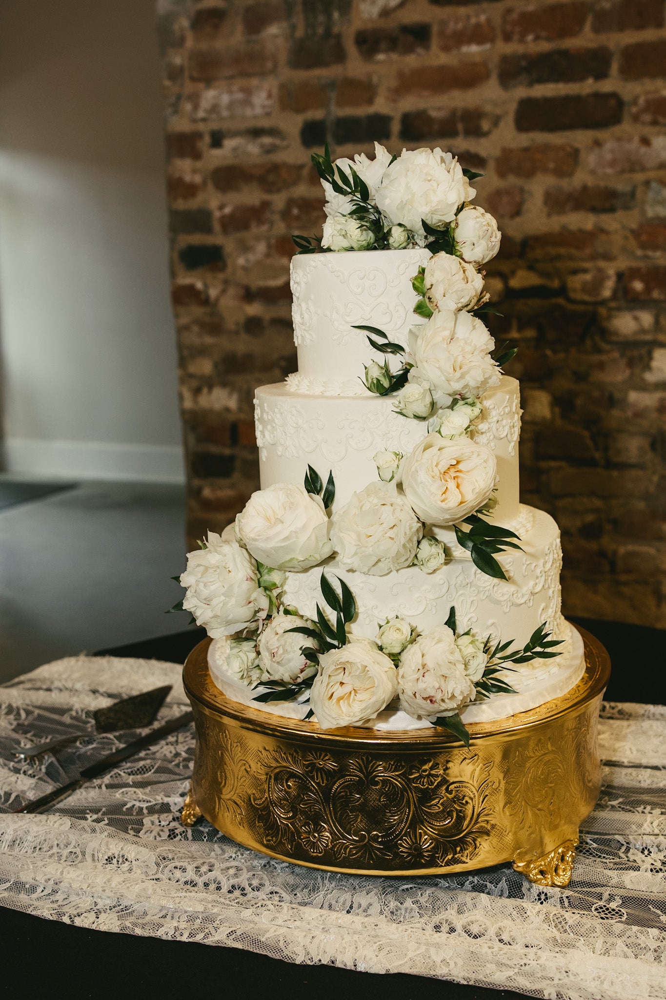 Multi-tiered wedding cake with floral decorations on a gold stand against a brick wall.