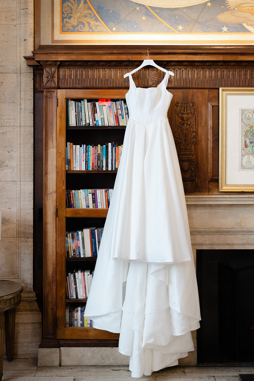 Galia Lahav Dove gown hanging on a wooden hanger against a decorative wall with books and artwork.