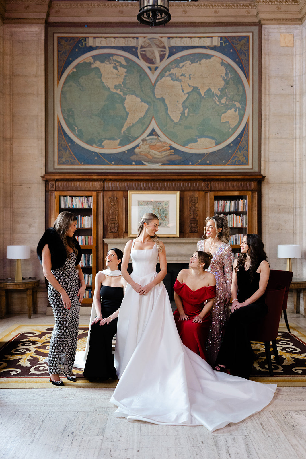 GL Bride Eliza in our Dove gown, standing with her bridesmaids in a room with a large world map on the wall