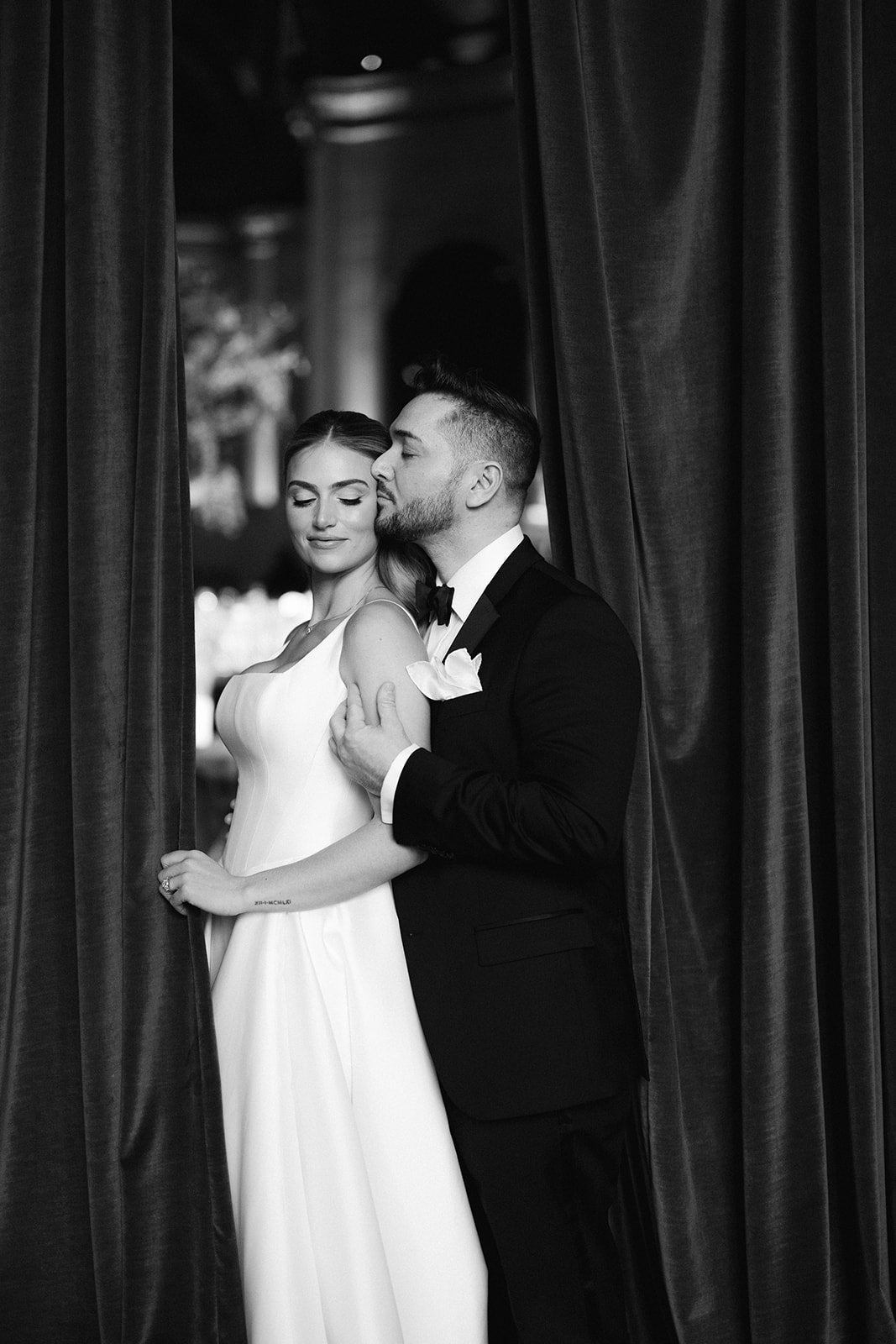 Black and white photo GL Bride Eliza and her husband embracing in front of draped curtains.