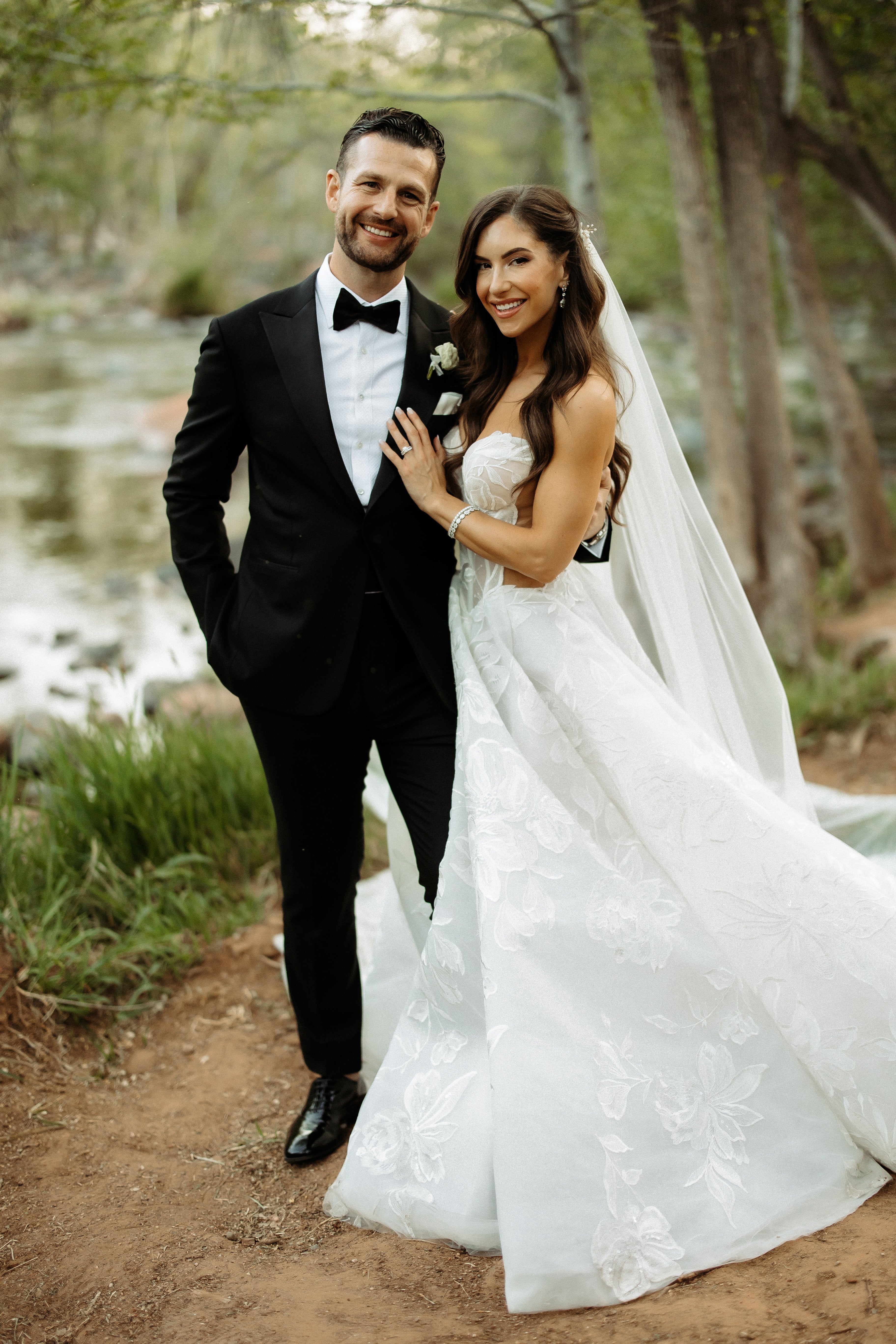 Man in a tuxedo and woman in a white wedding dress standing in a forest setting.