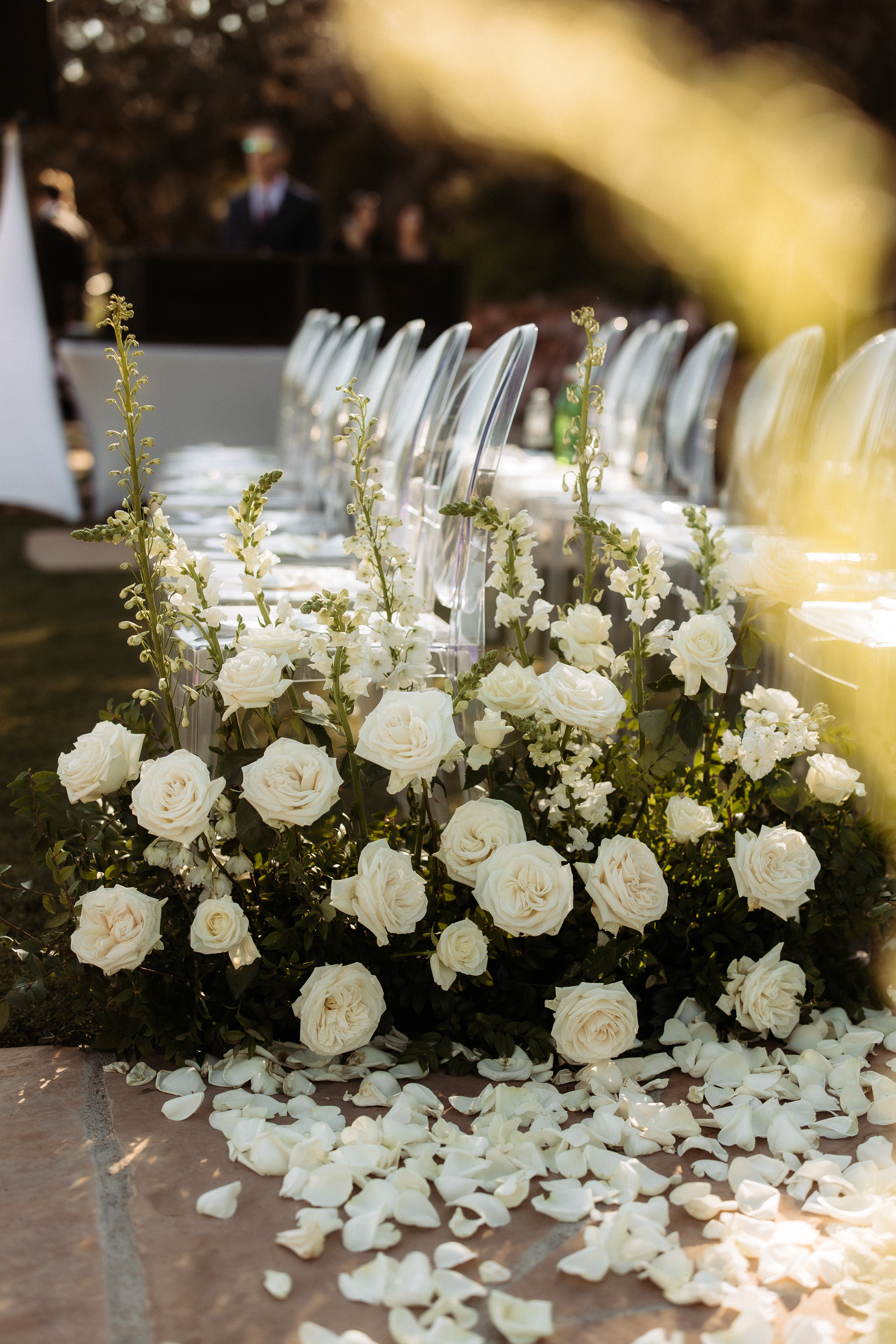 Decorative floral arrangement with white flowers and greenery on a table with clear plastic chairs.