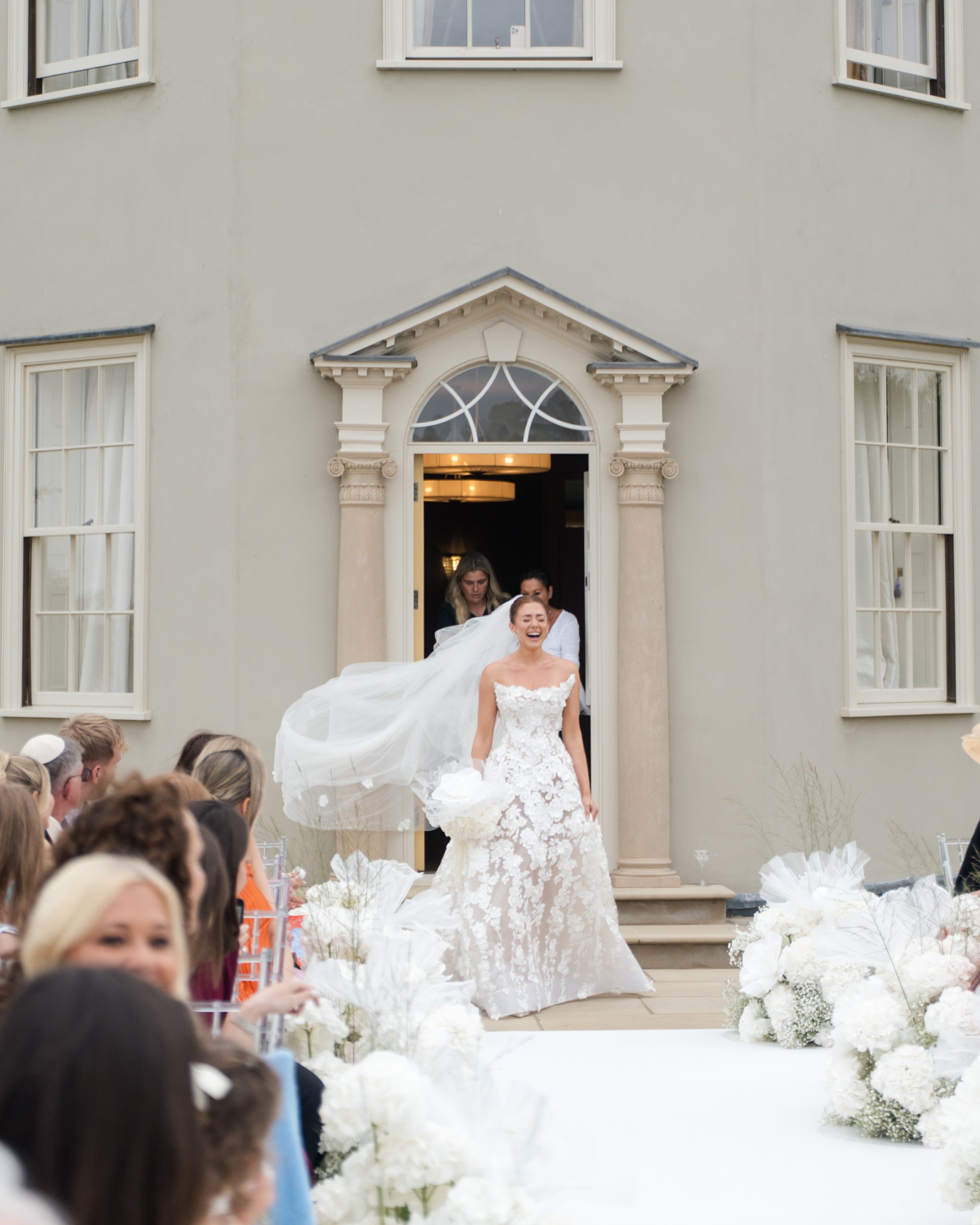 GL Bride Emily in our Rosemary gown walking out of a grand building with guests and floral decorations.
