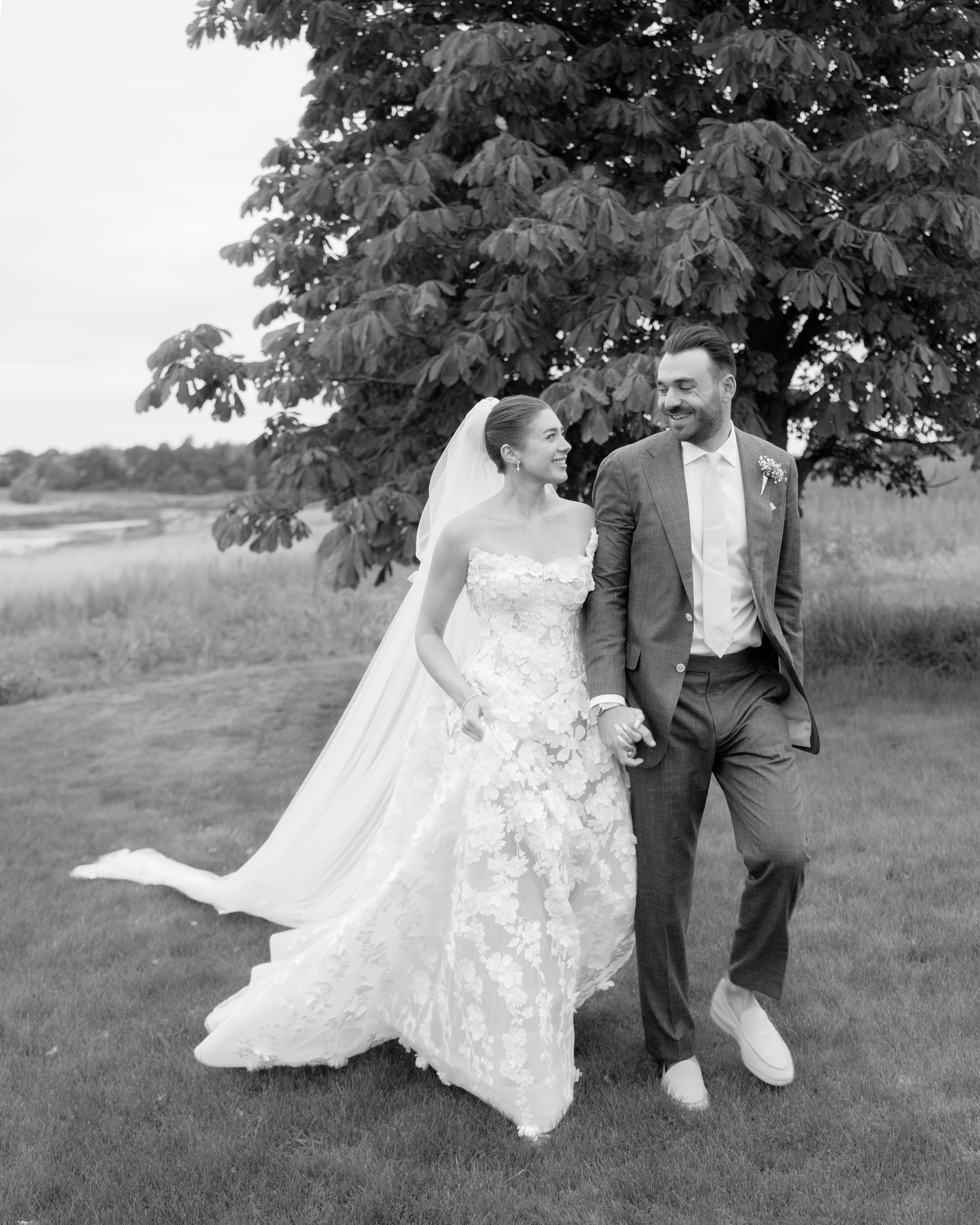 Black and white photo of GL Bride Emily in our Rosemary and groom walking outdoors with trees in the background.
