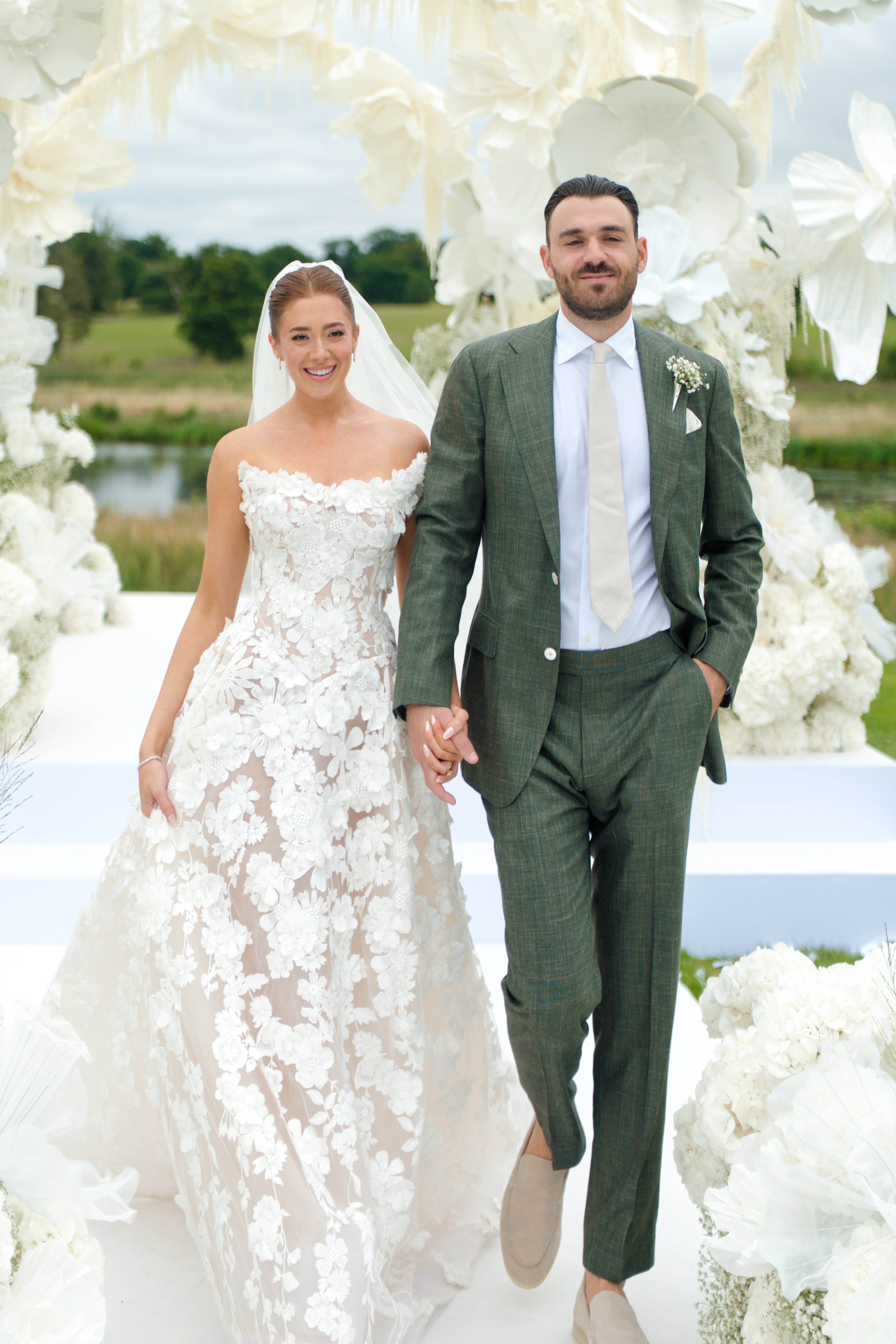 GL Bride Emily in our Rosemary gown and husband standing under a floral archway.