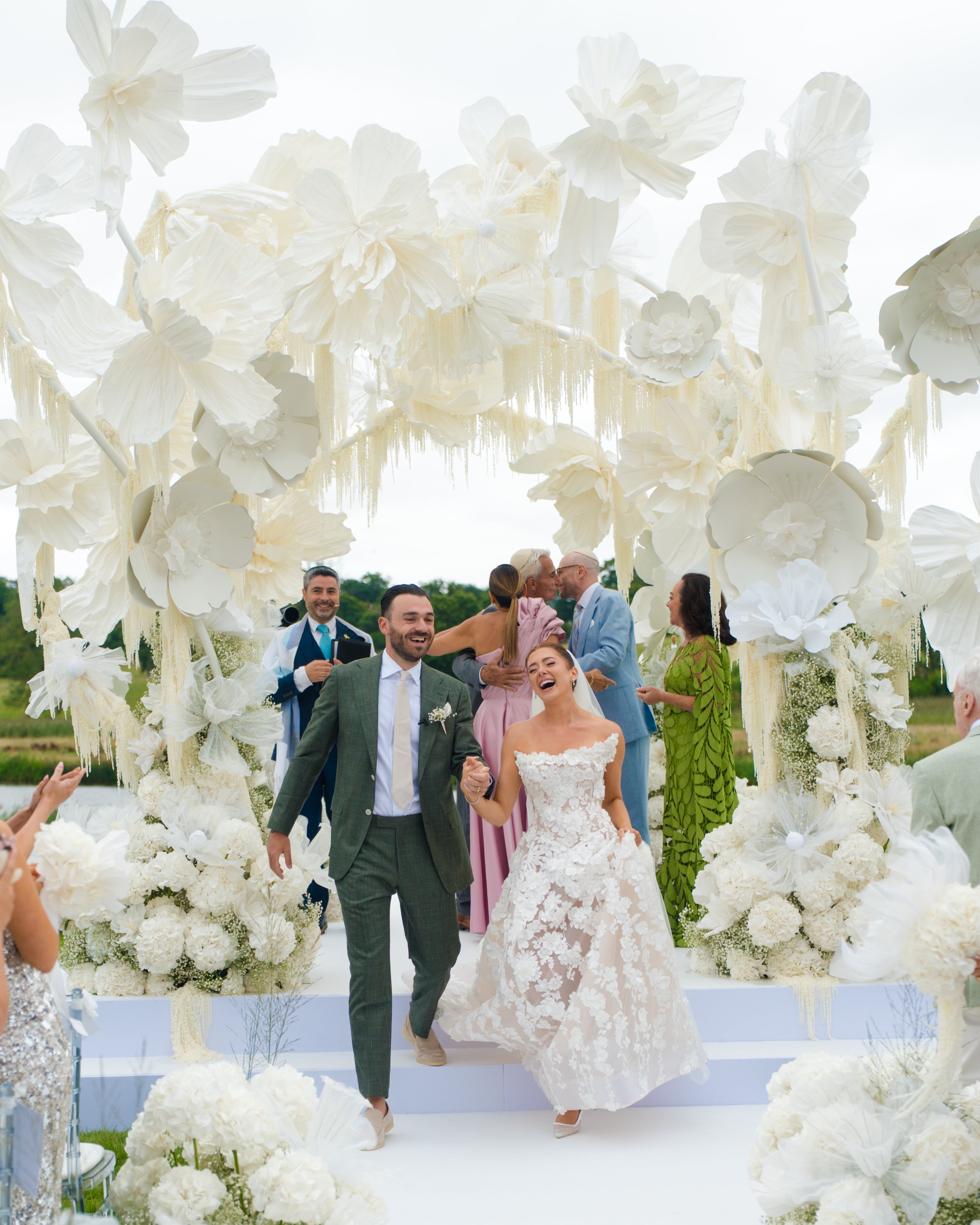 Wedding couple walking under a large floral arch with guests in the background.