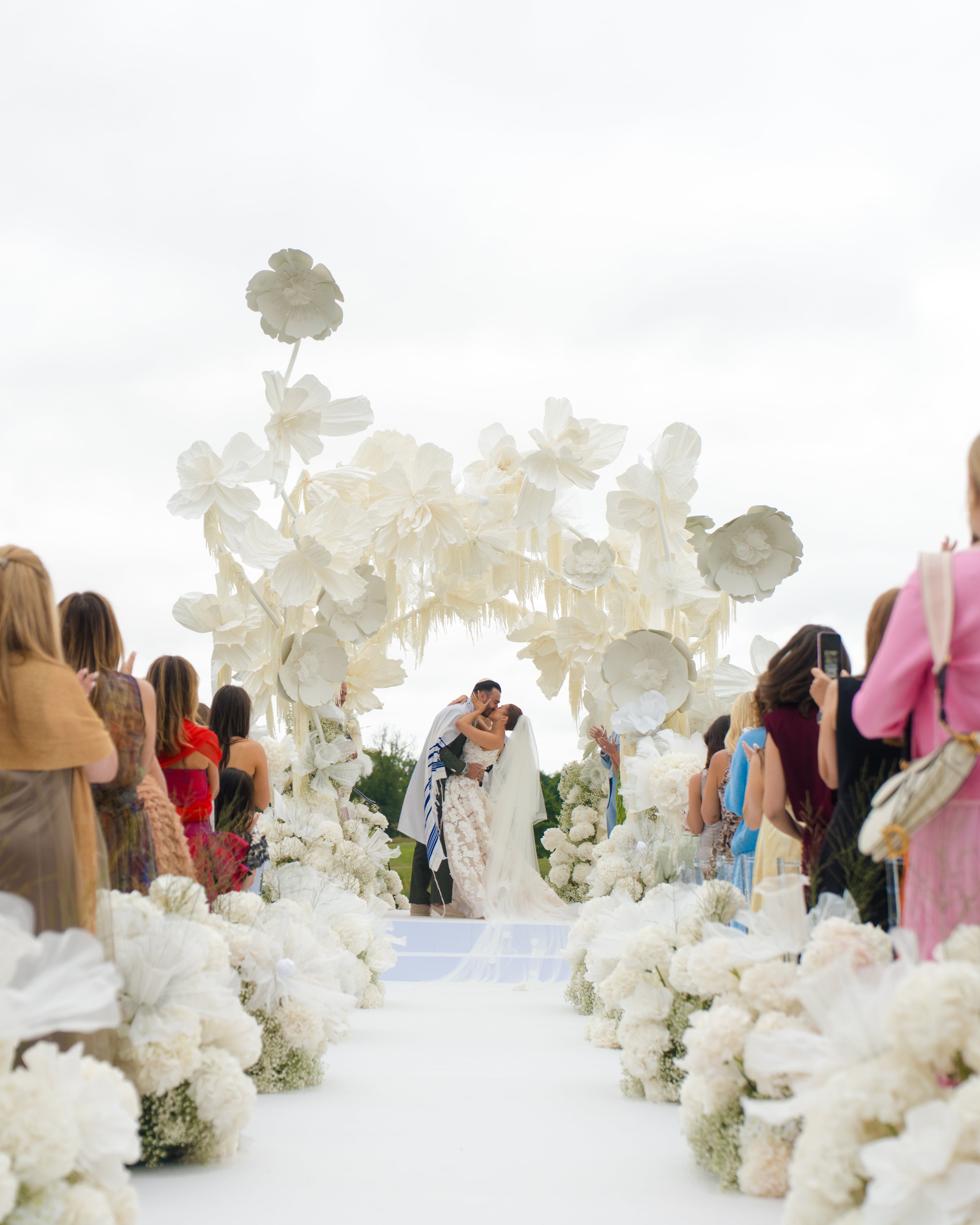 GL Bride Emily in our Rosemary kissing her husband under a floral arch with guests on either side.