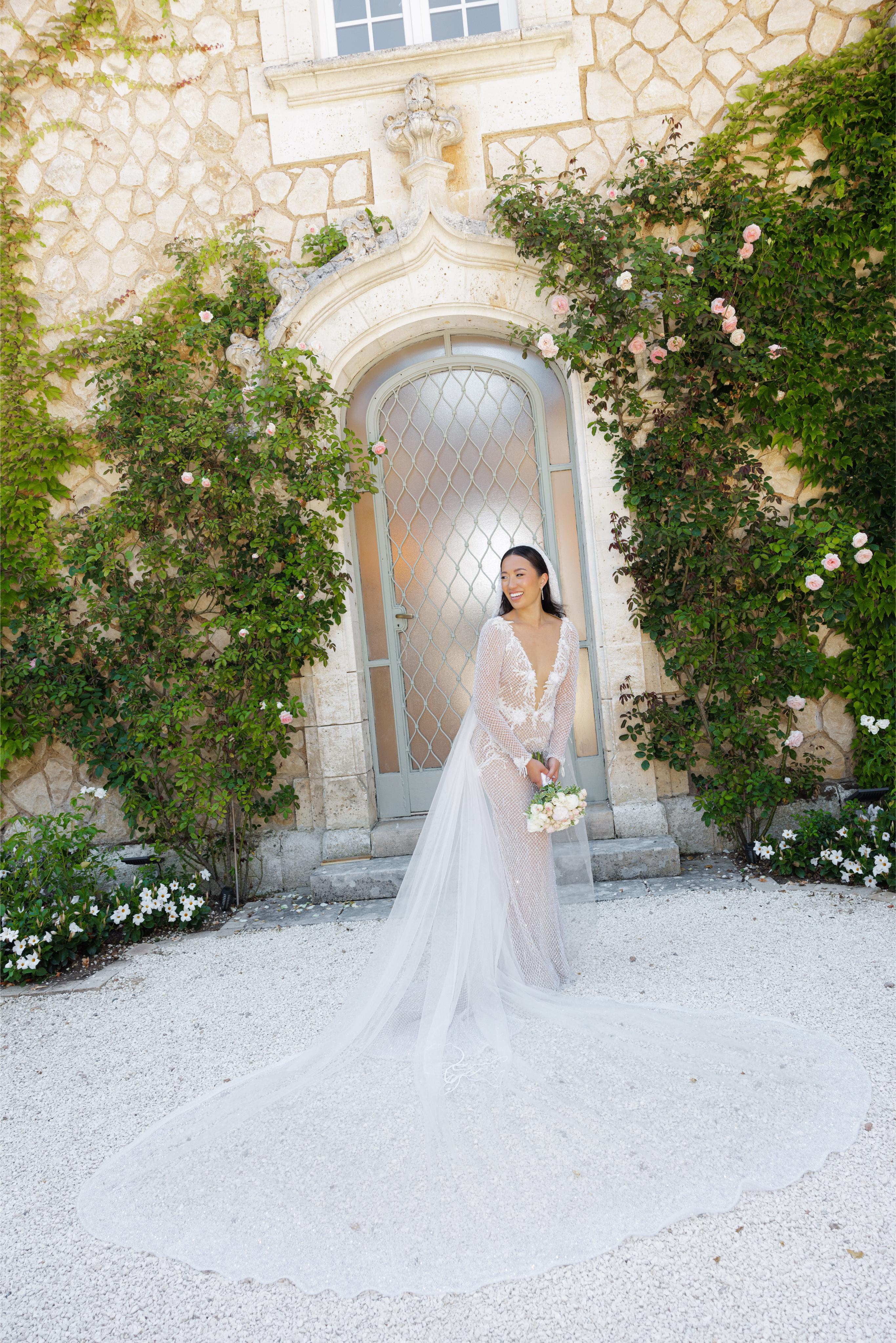 Woman in a white wedding dress standing in front of an elegant stone building with greenery.