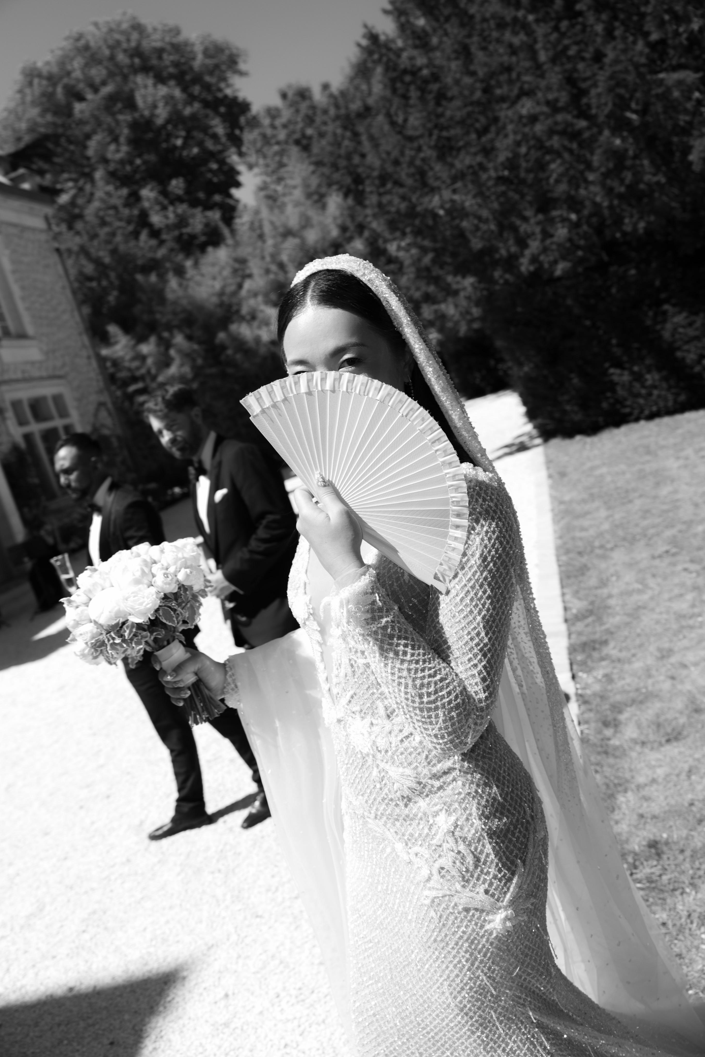 Black and white photo of a woman in a wedding dress holding a fan outdoors.