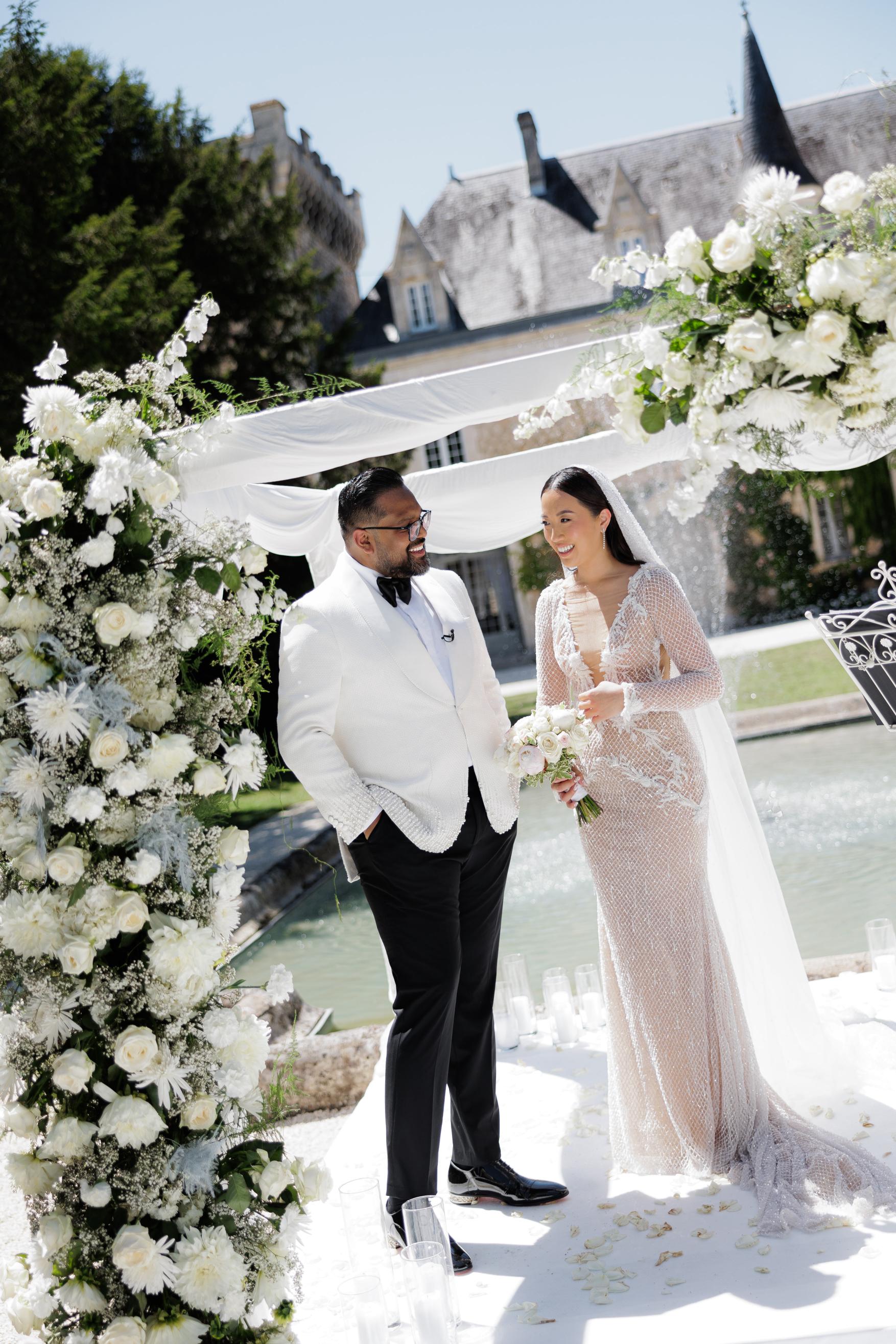 Wedding couple standing under a floral archway with a building in the background.