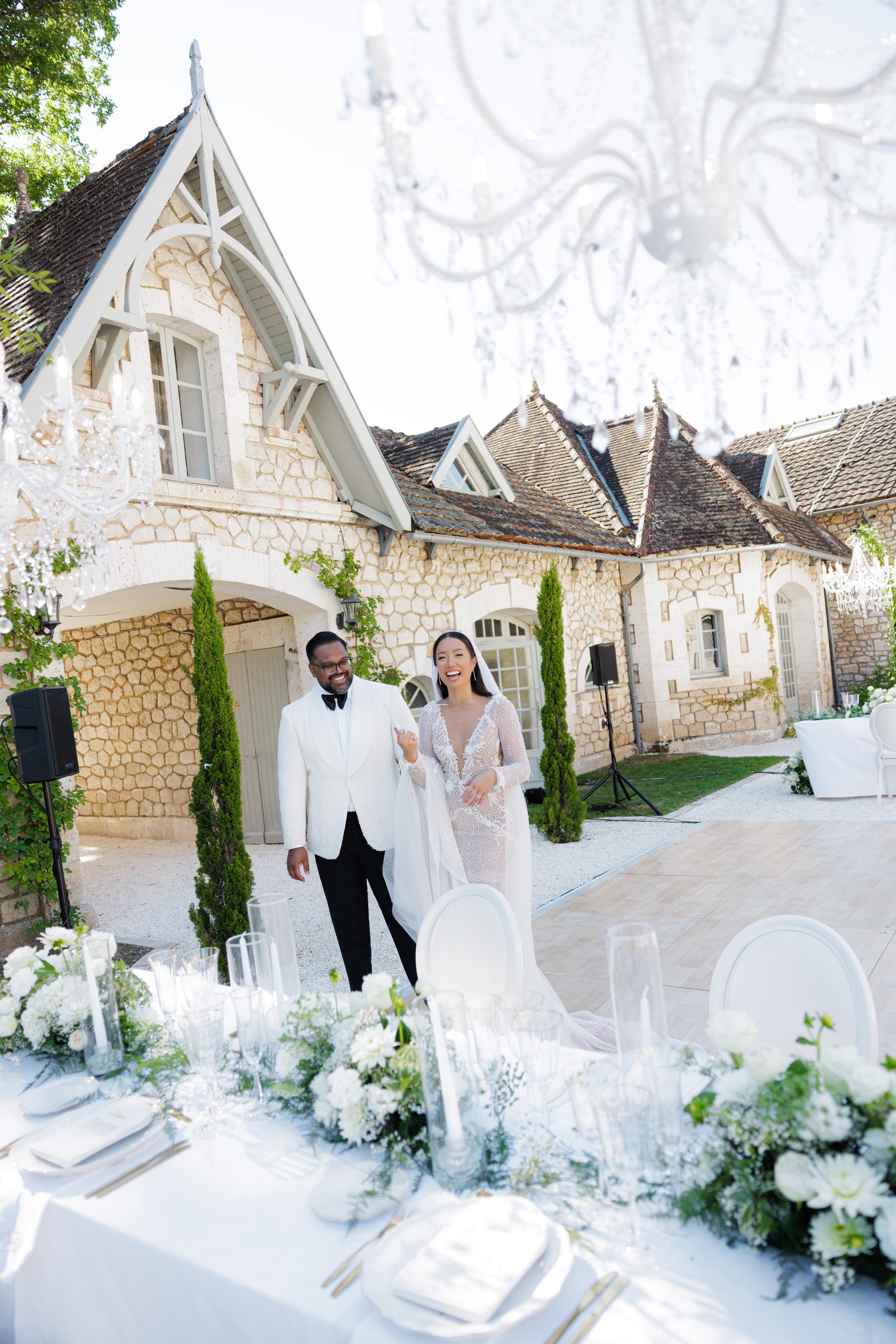 Wedding couple standing in front of a stone building with white floral arrangements and chairs.
