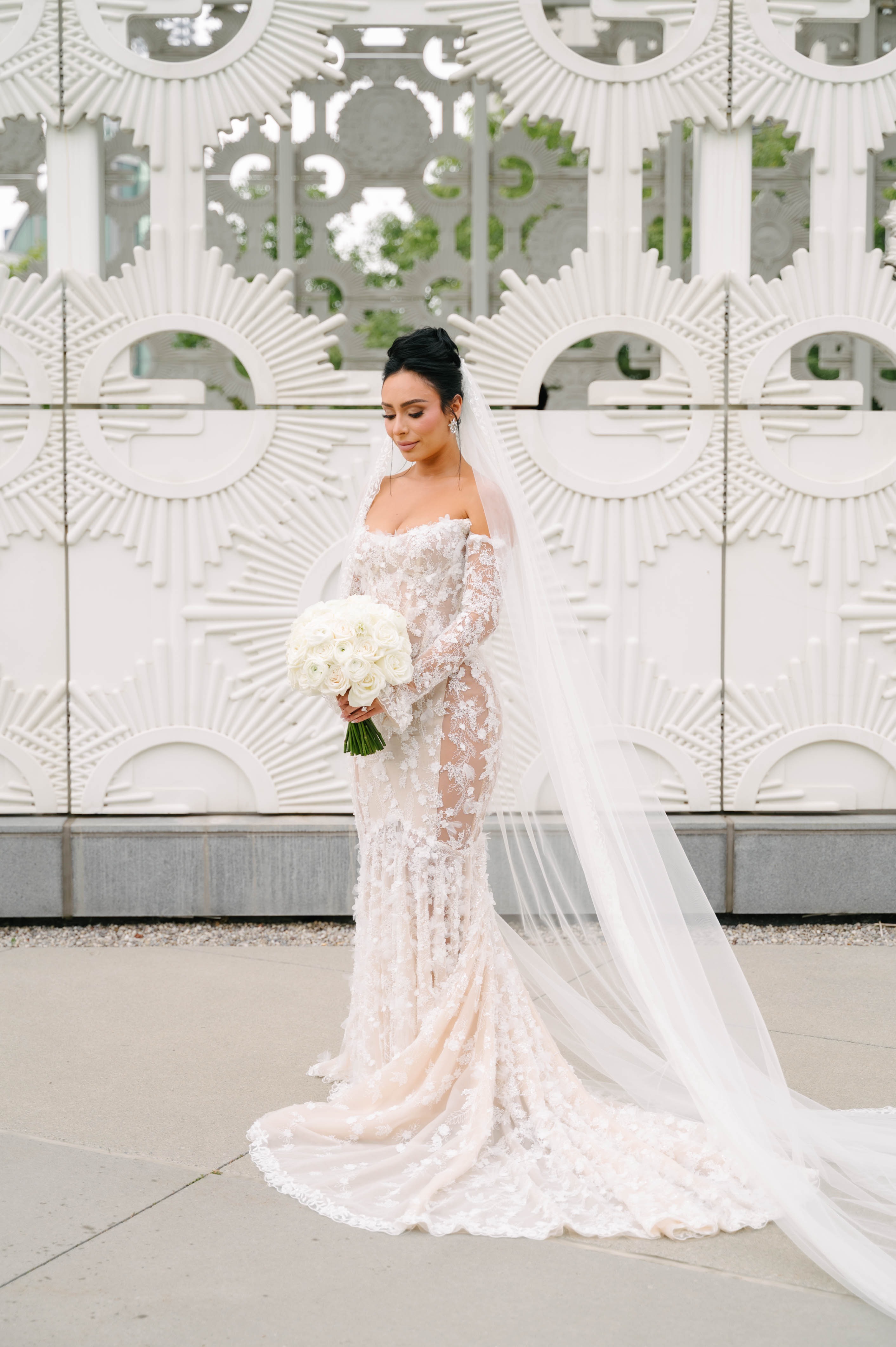 GL Bride Eva in our Maya dress with a long train standing in front of a decorative white wall.
