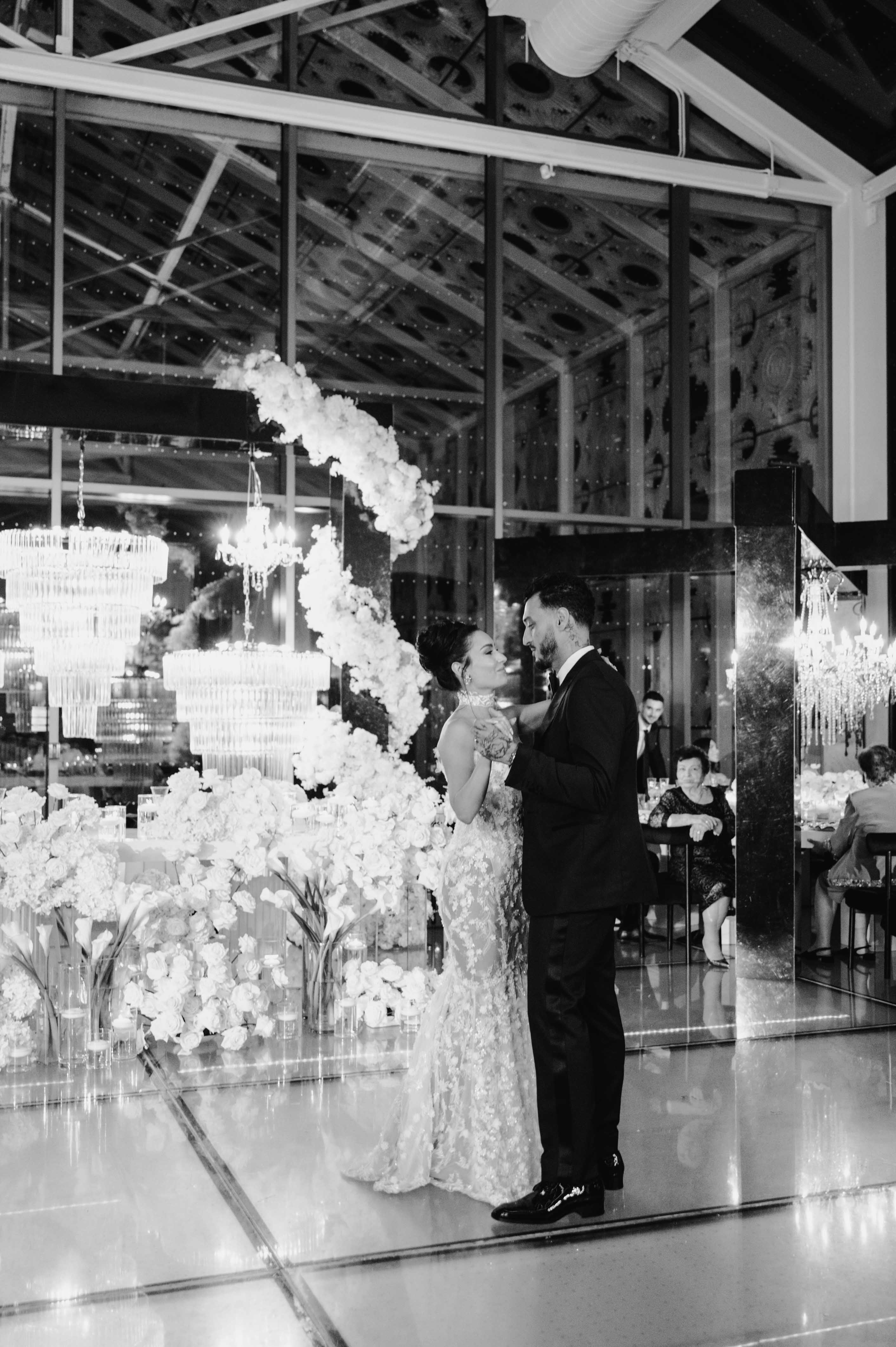Black and white photo of GL Bride Eva in our Maya gown and husband  in formal attire in a decorated room with floral arrangements.