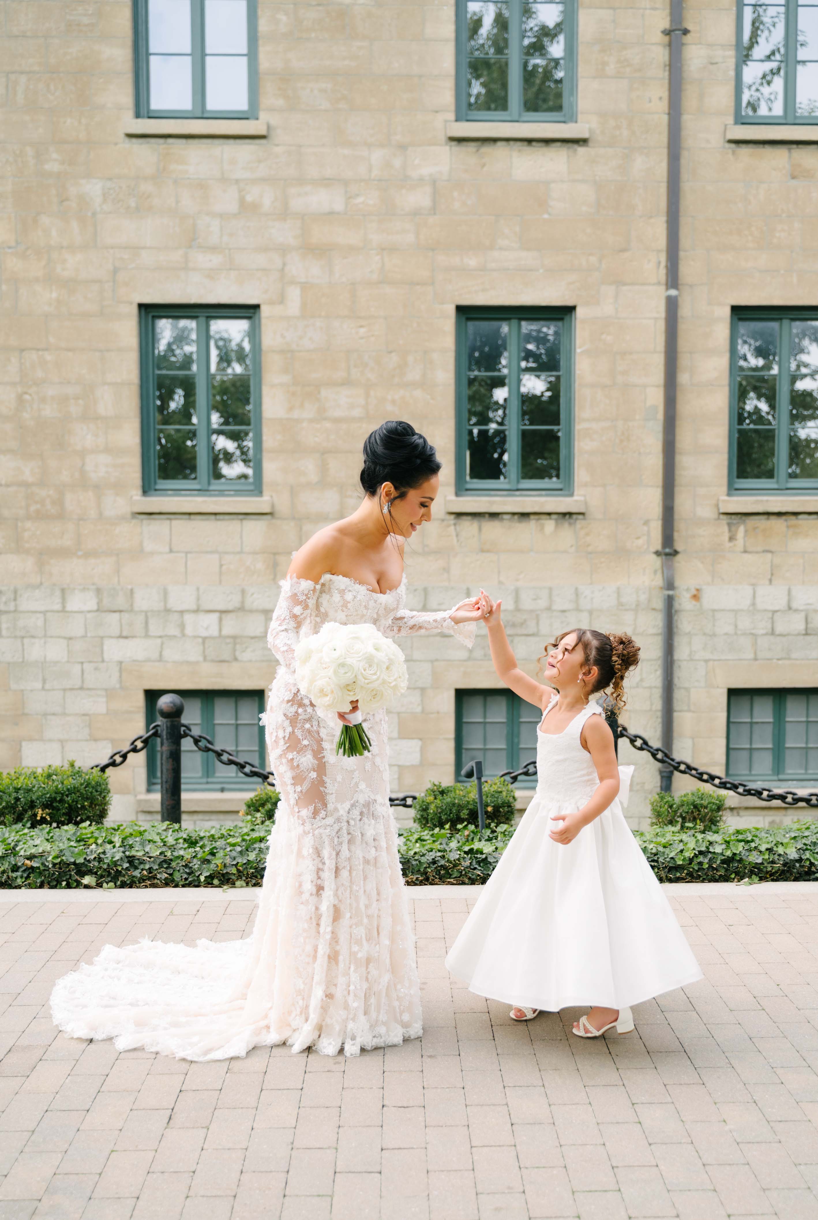 GL Bride Eva in our Maya gown holding hands with a young girl in a white dress in front of a brick building.