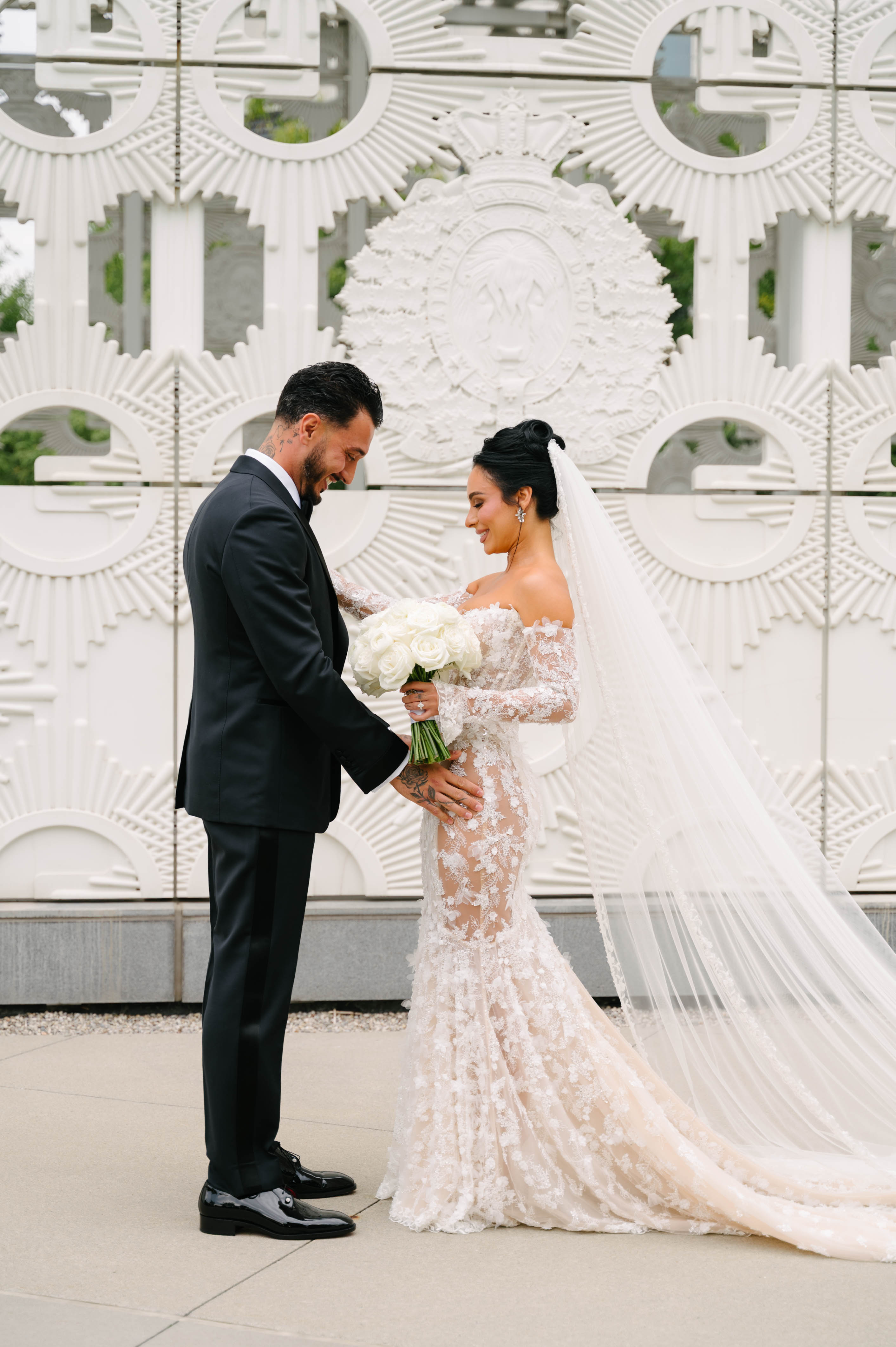 GL Bride Eva in a custom Maya dress  with her husband in front of a decorative white wall.
