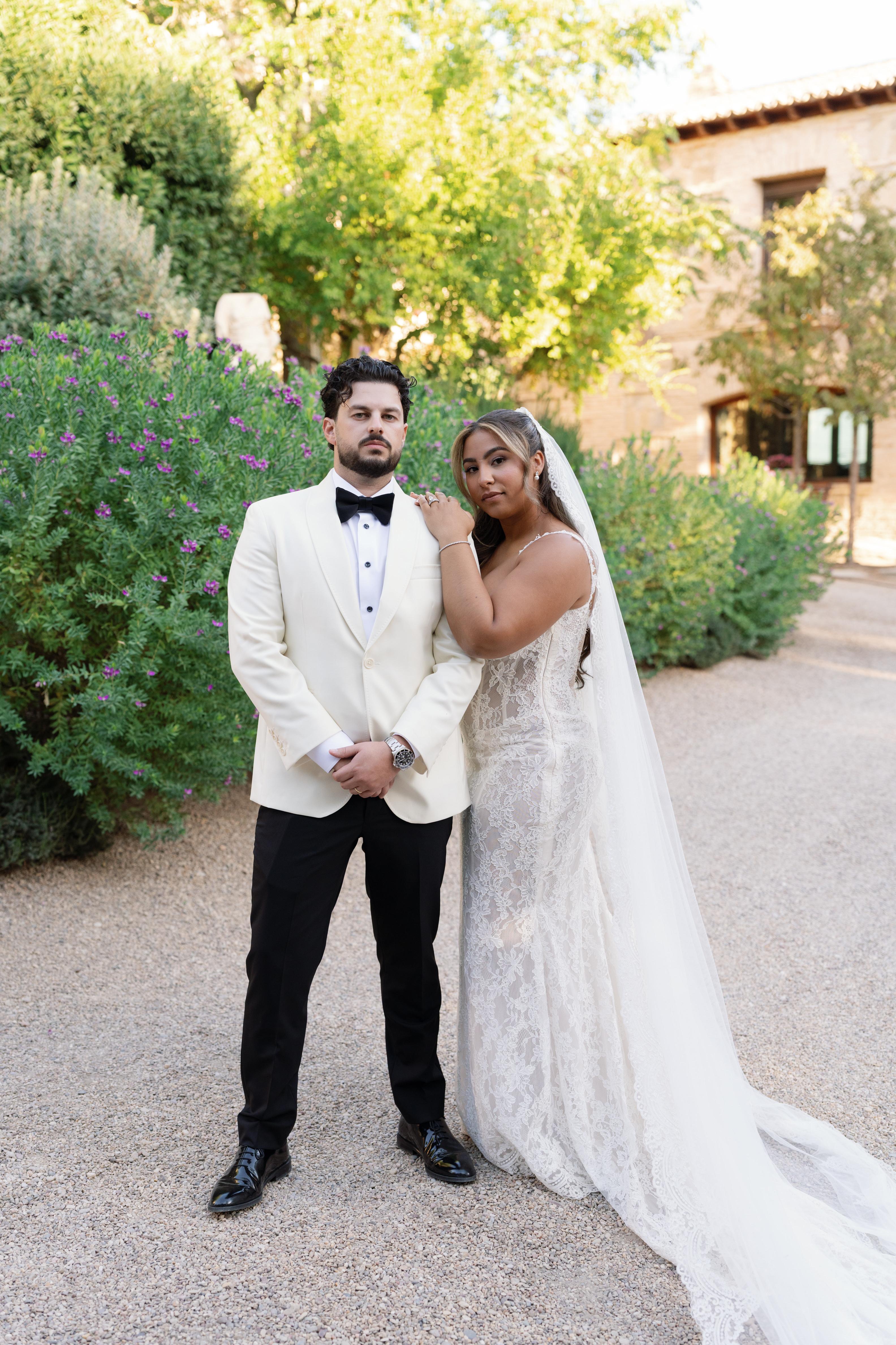 Man in a white suit and bow tie, GL Bride Janai in our Anais with a long veil, standing outdoors.