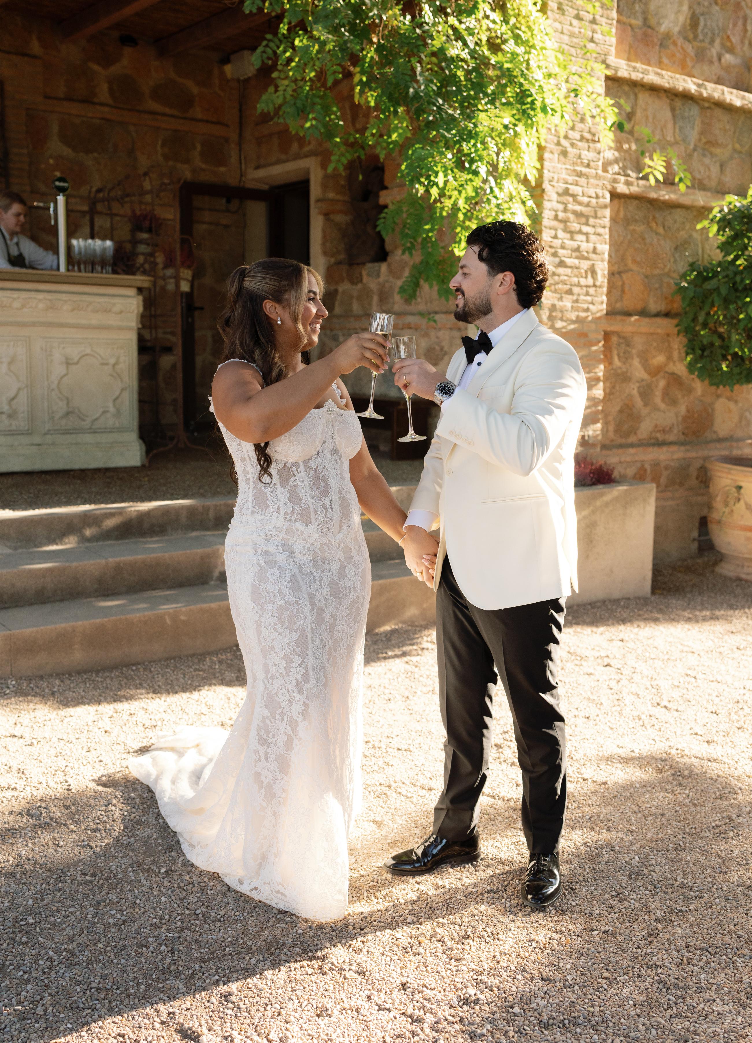 GL Bride Janai in our Anais with her husband in formal attire standing outdoors with a stone building in the background