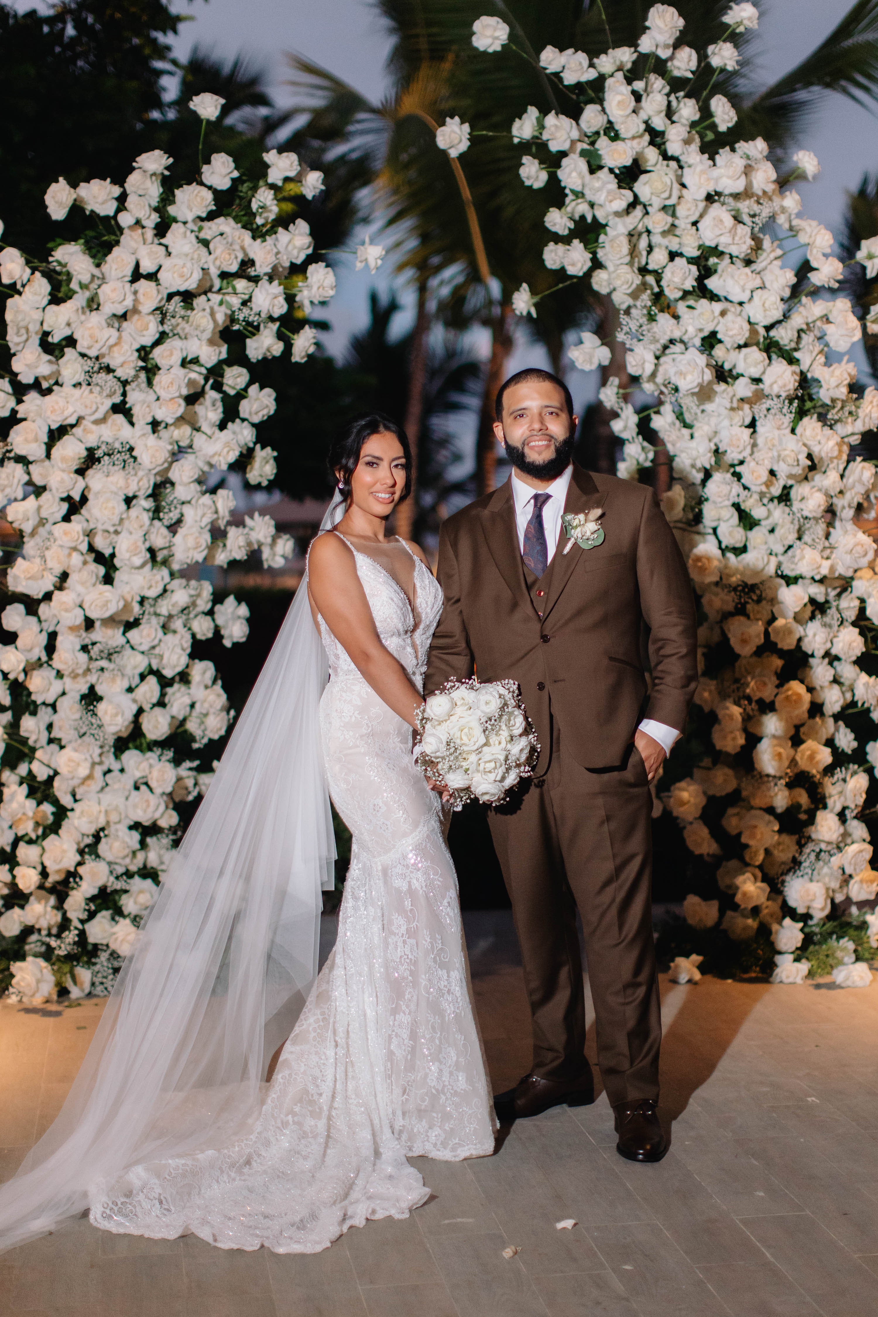 Wedding couple standing in front of a floral archway with white flowers. GL Bride Jennifer wearing our G-404