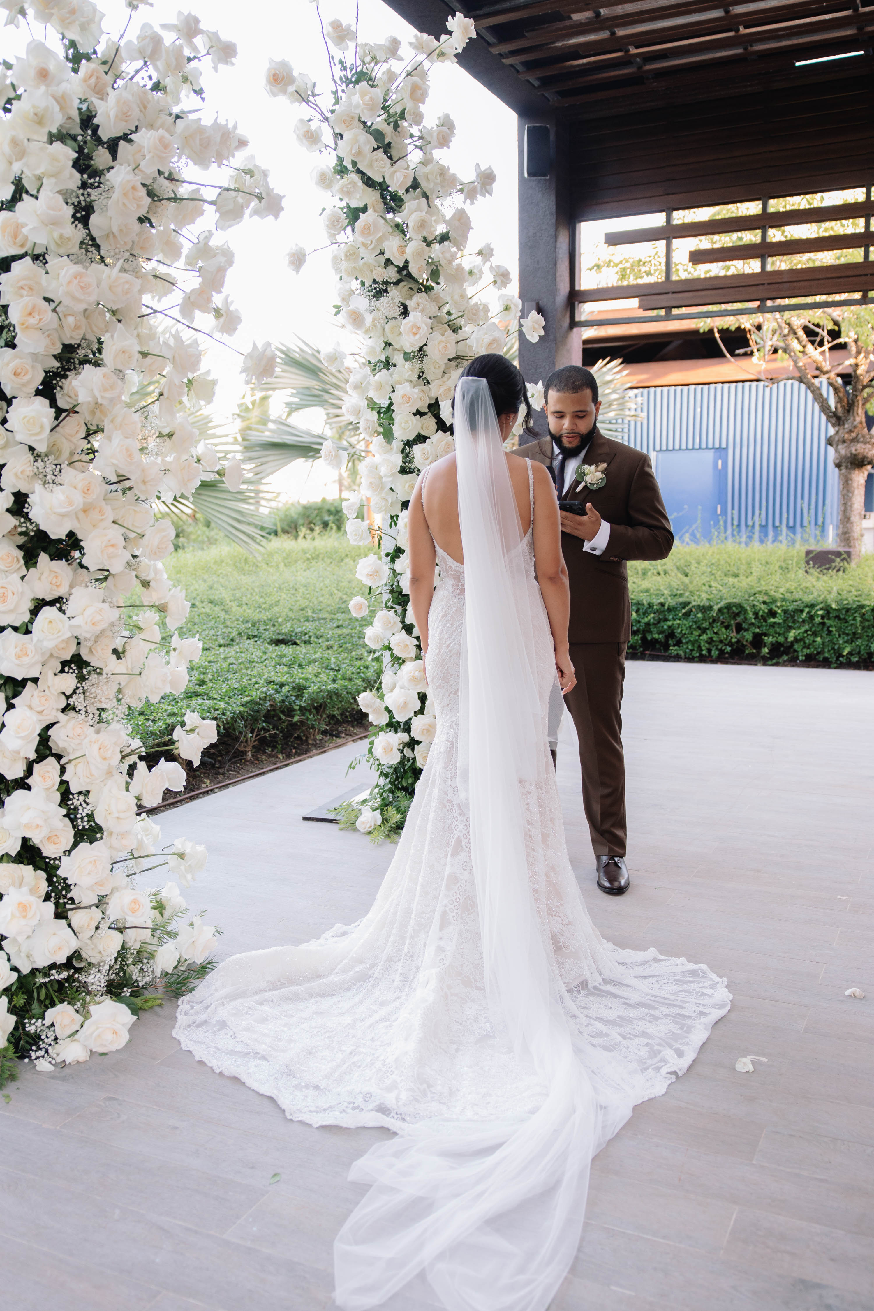 Wedding couple standing under a floral archway with a modern building in the background. GL Bride Jennifer wearing our G-404
