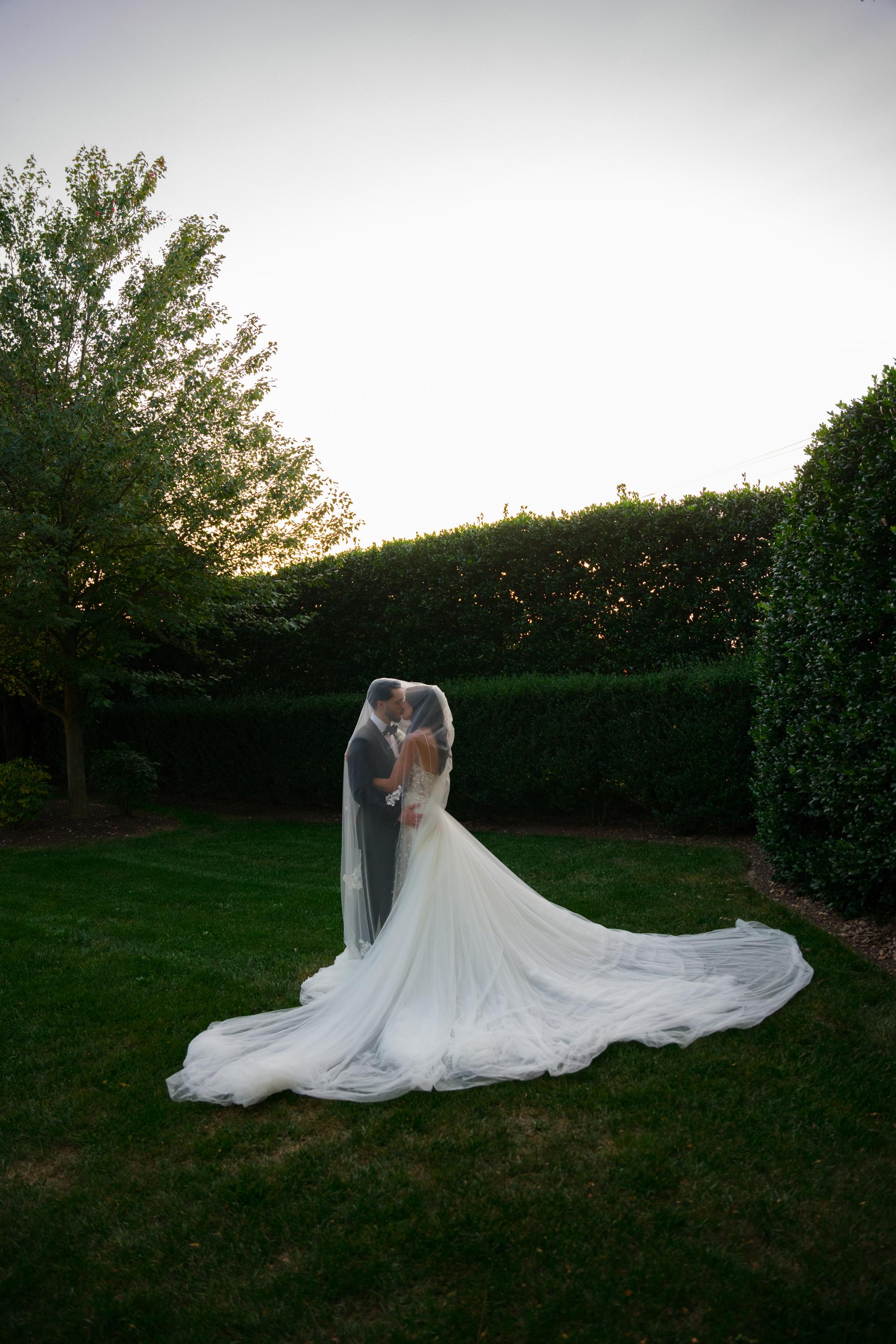 GL Bride Jessica wearing our Catalina gown and her husband embracing in a garden setting with a long veil.