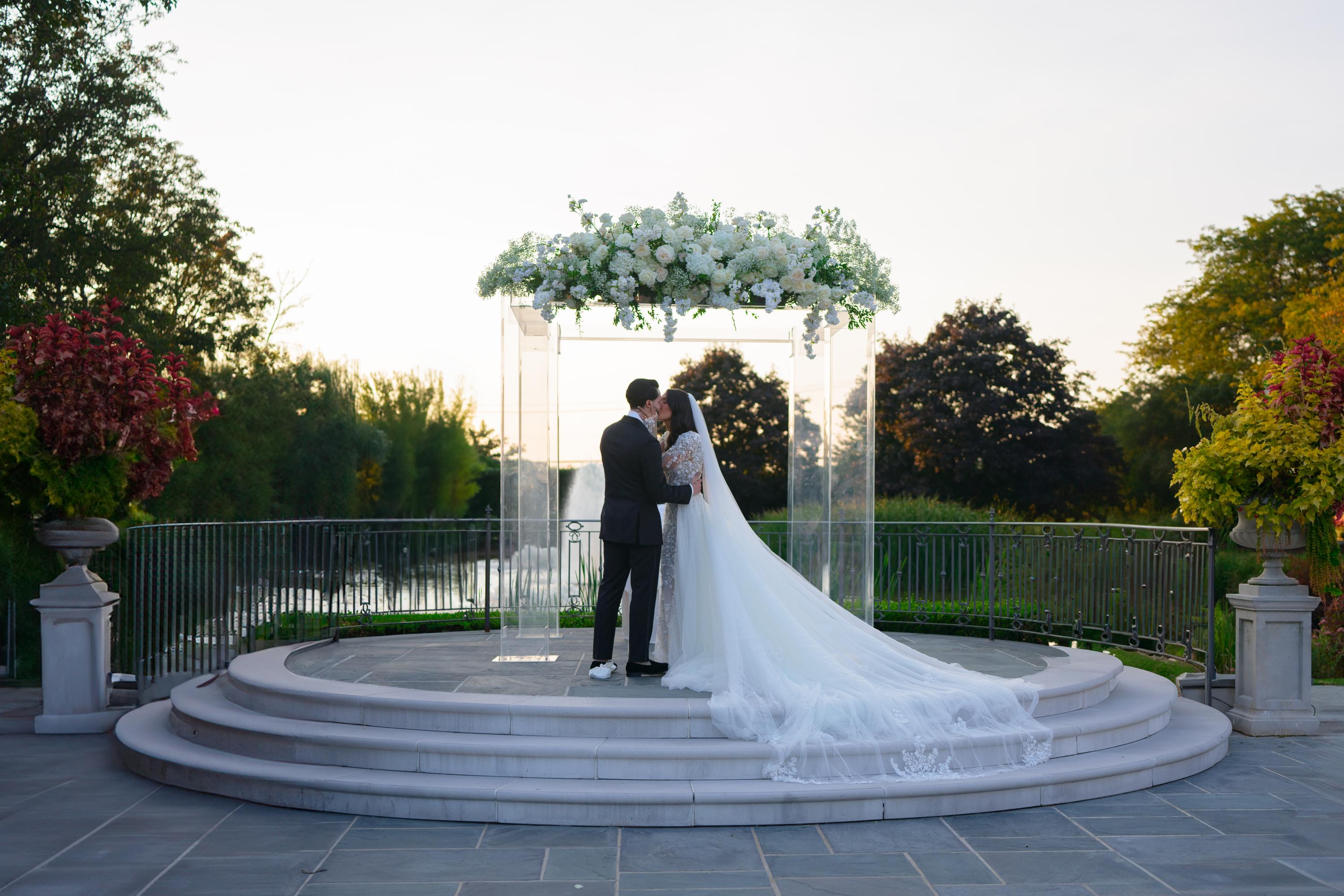 GL Bride Jessica wearing our Catalina gown and  her husband standing under a floral archway with a scenic background