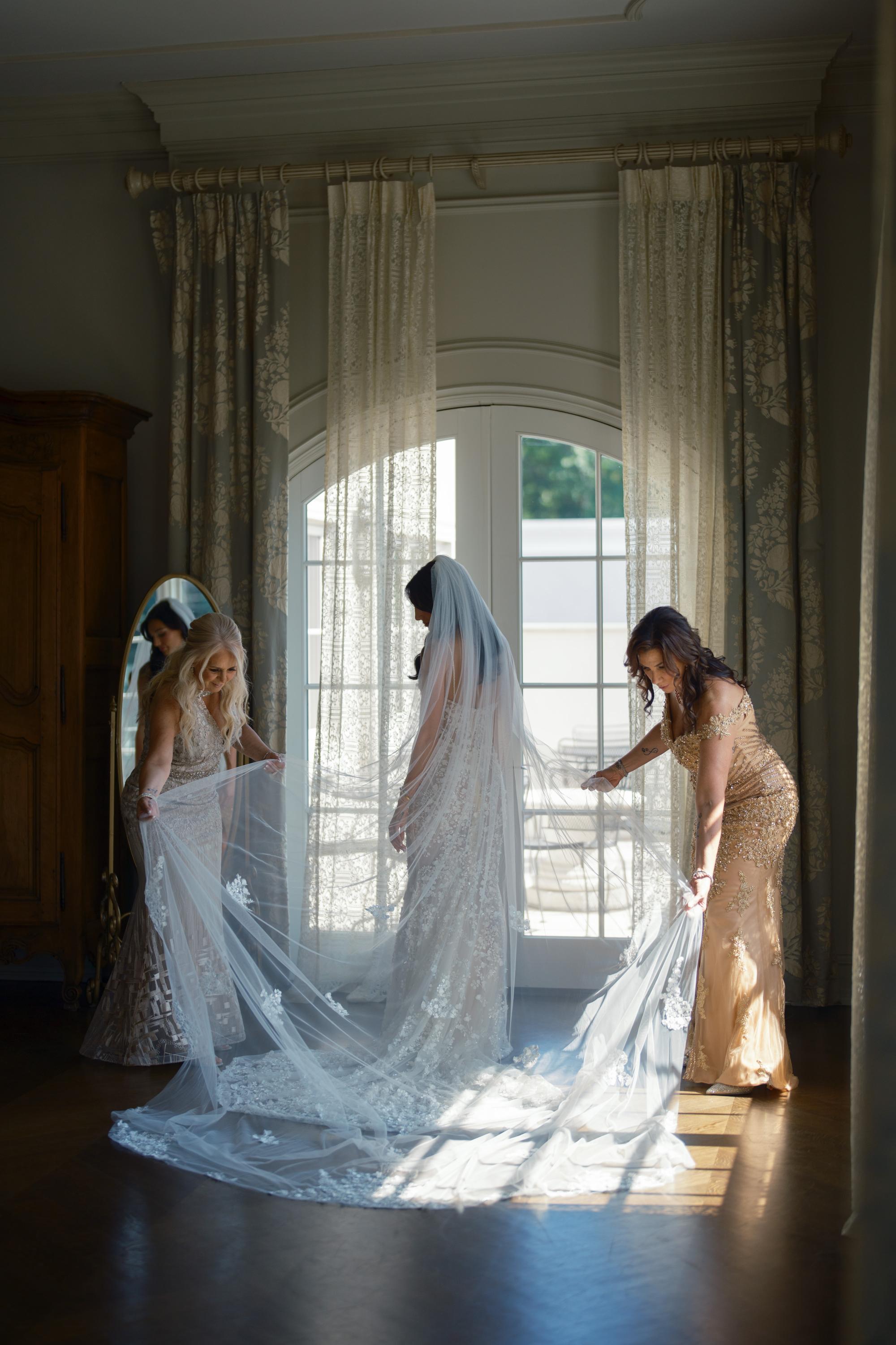 Bridesmaid in a gold dress assisting our GL Bride Jessica with Galia Lahav Catalina  Gown by a large window.