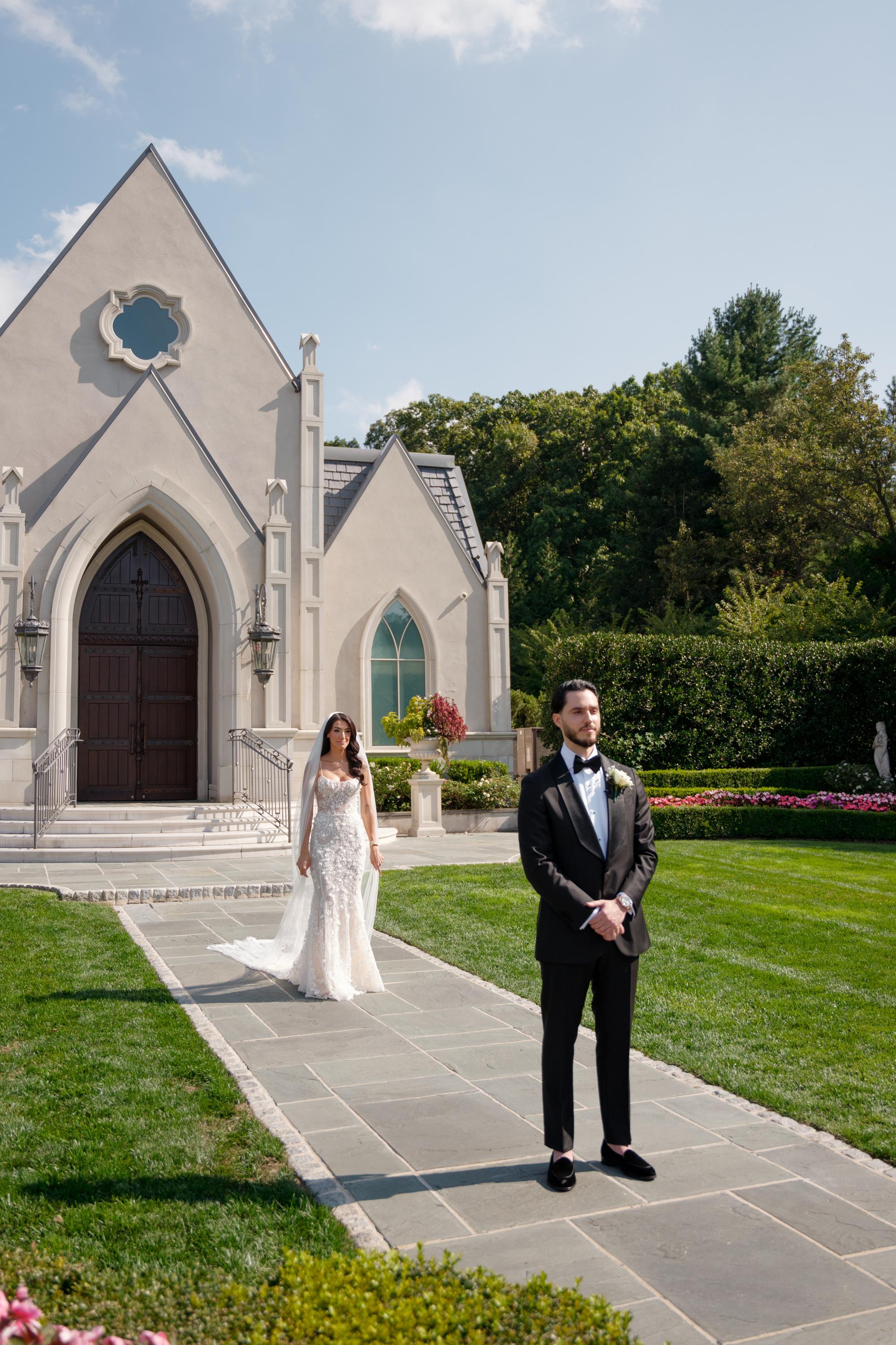 GL Bride Jessica wearing our Catalina gown with her husband, standing in front of a chapel with greenery and flowers.