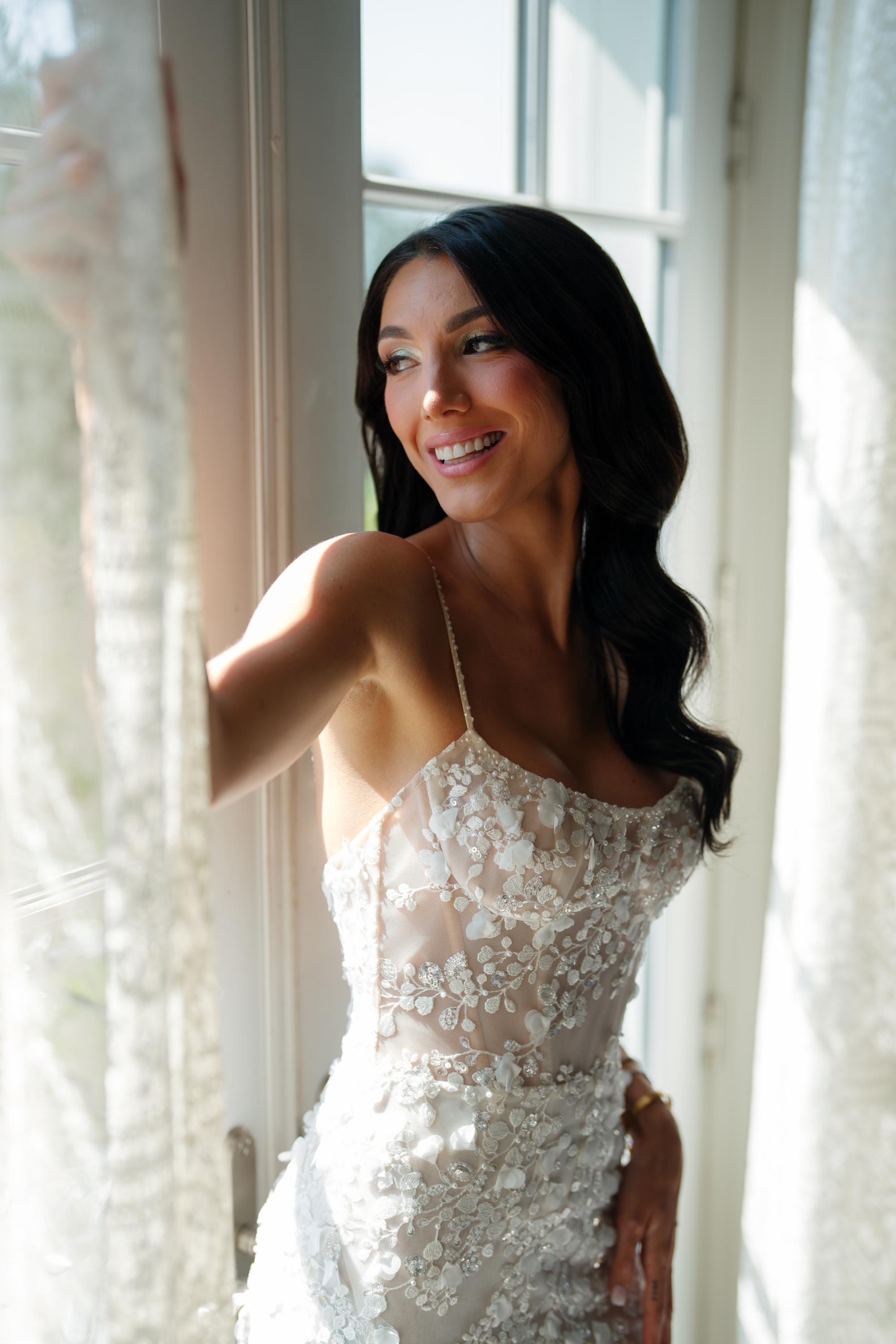 GL Bride Jessica wearing our Catalina gown standing in a bright room with large windows.