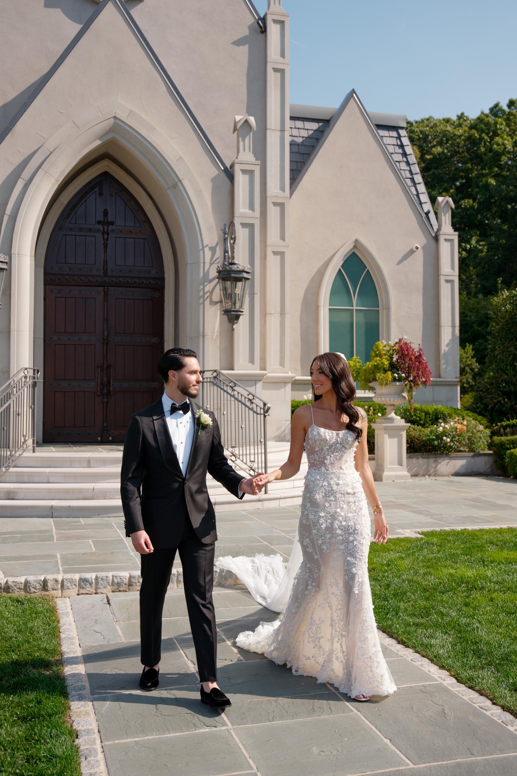 GL Bride Jessica wearing our Catalina gown  and her husband walking hand-in-hand in front of a church.