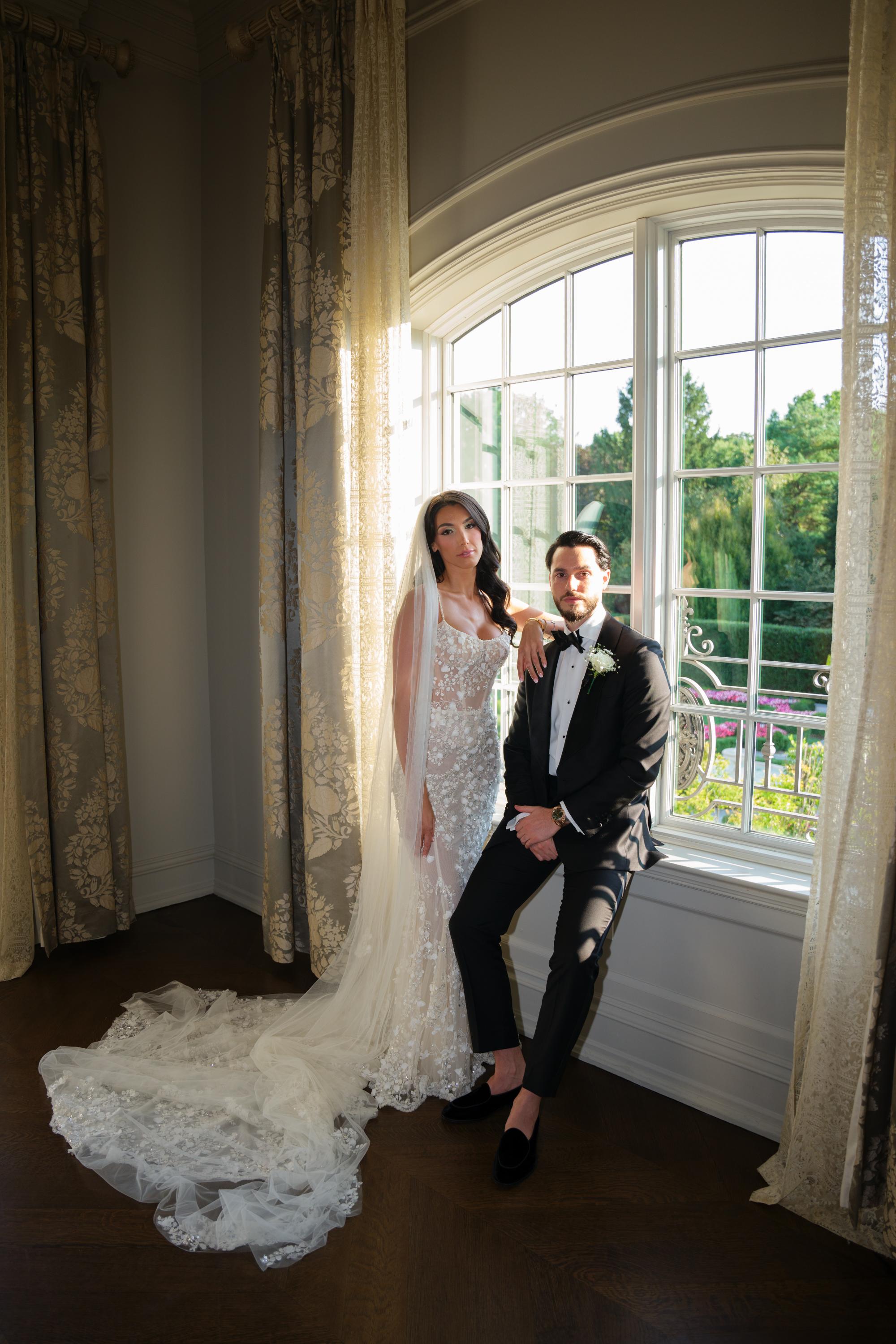 GL Bride Jessica wearing our Catalina gown and her husband in a room with large windows and decorative curtains.