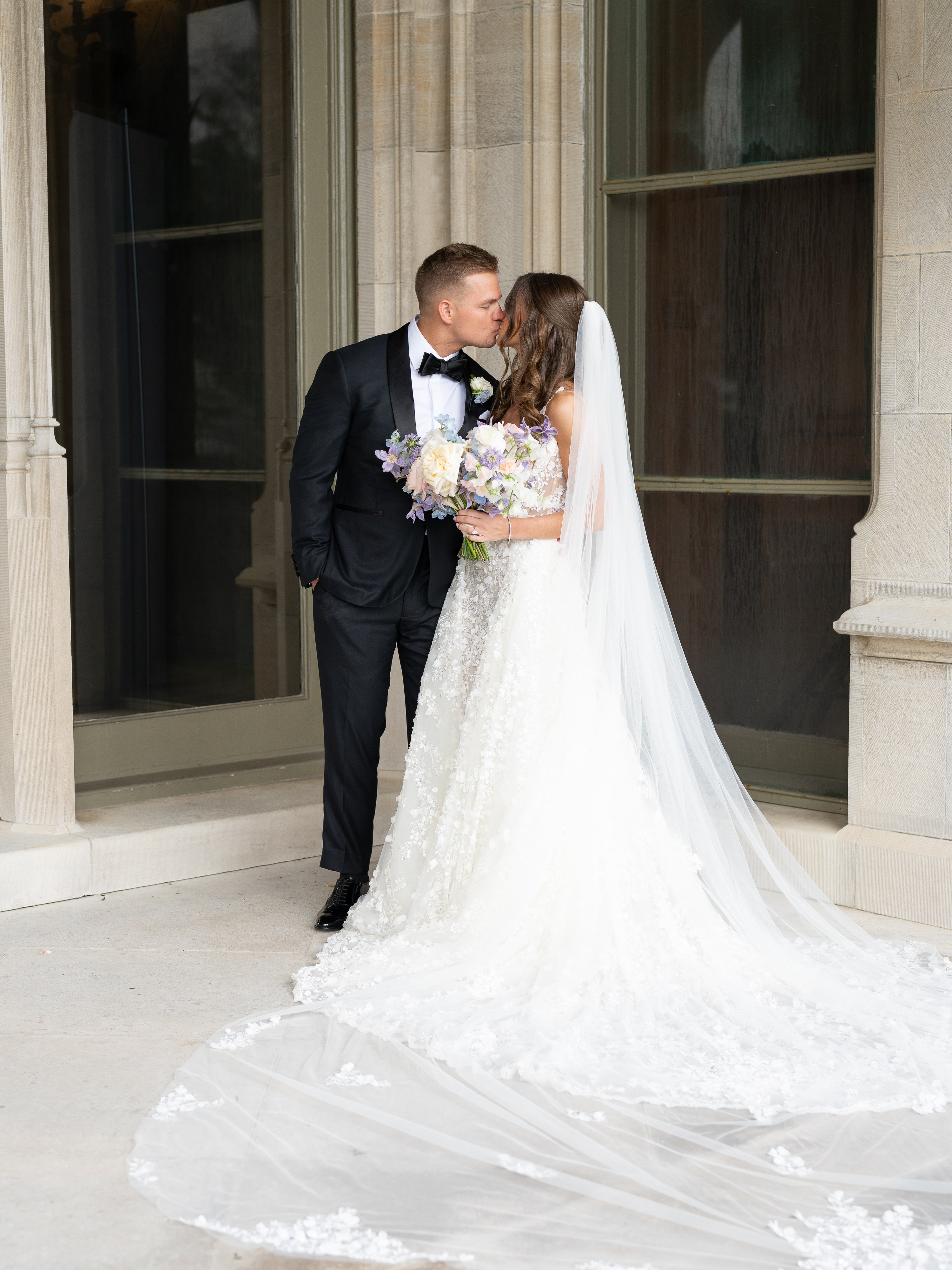 Man in a tuxedo and woman in a white wedding dress with a long veil standing in front of a classical building.