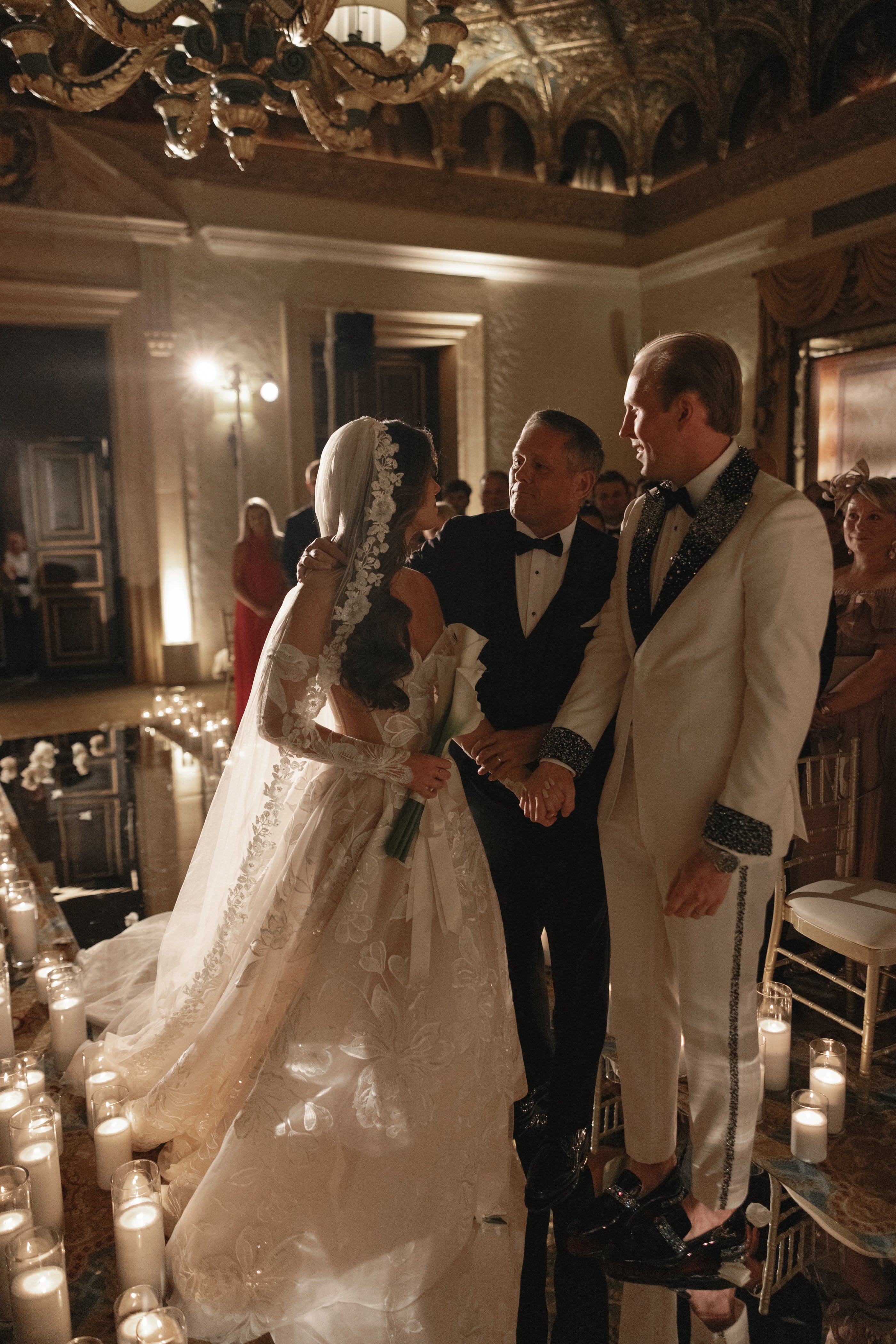 Wedding ceremony in a grand room with chandeliers and guests.