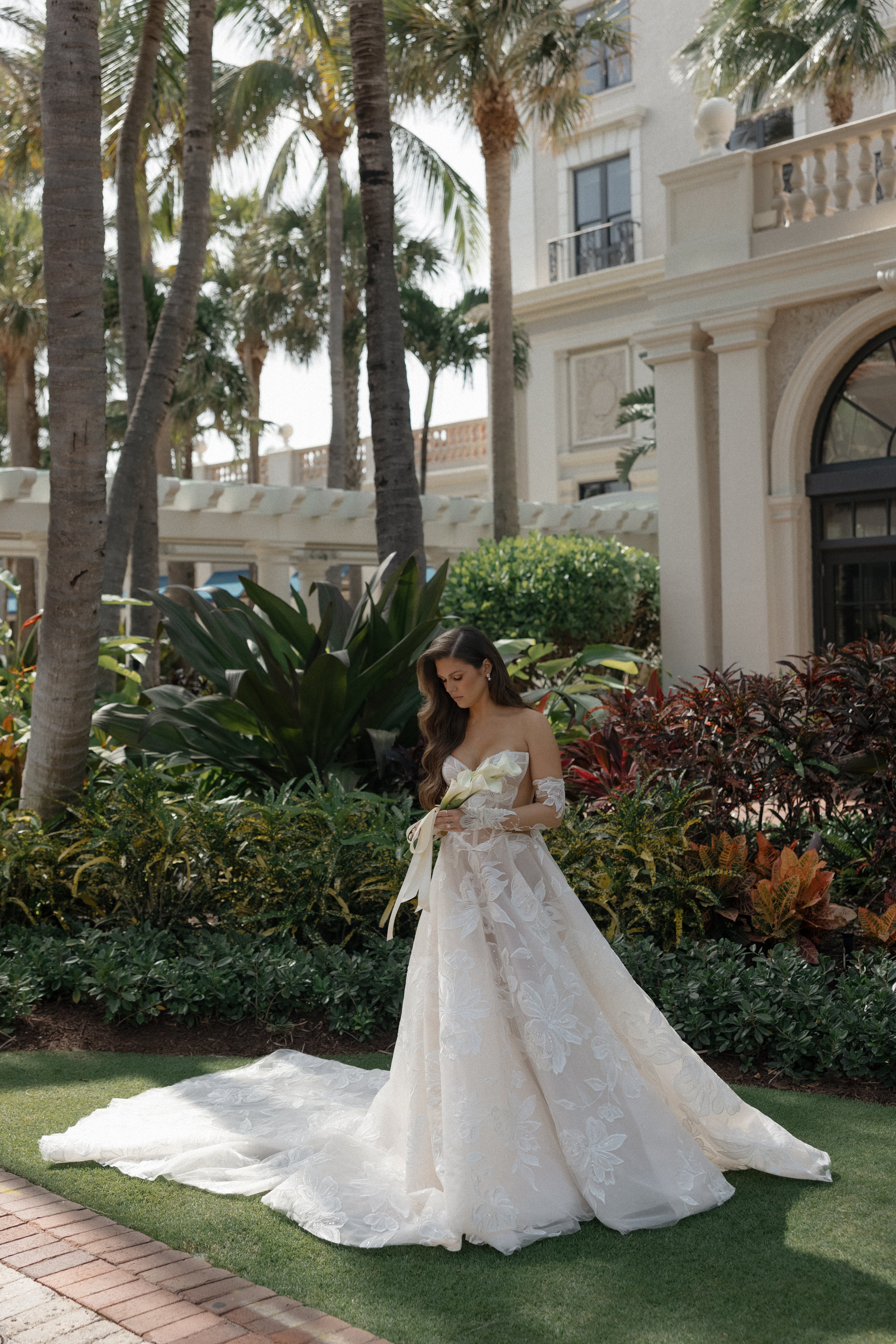 Woman in a white wedding dress standing in a garden with palm trees and a building in the background