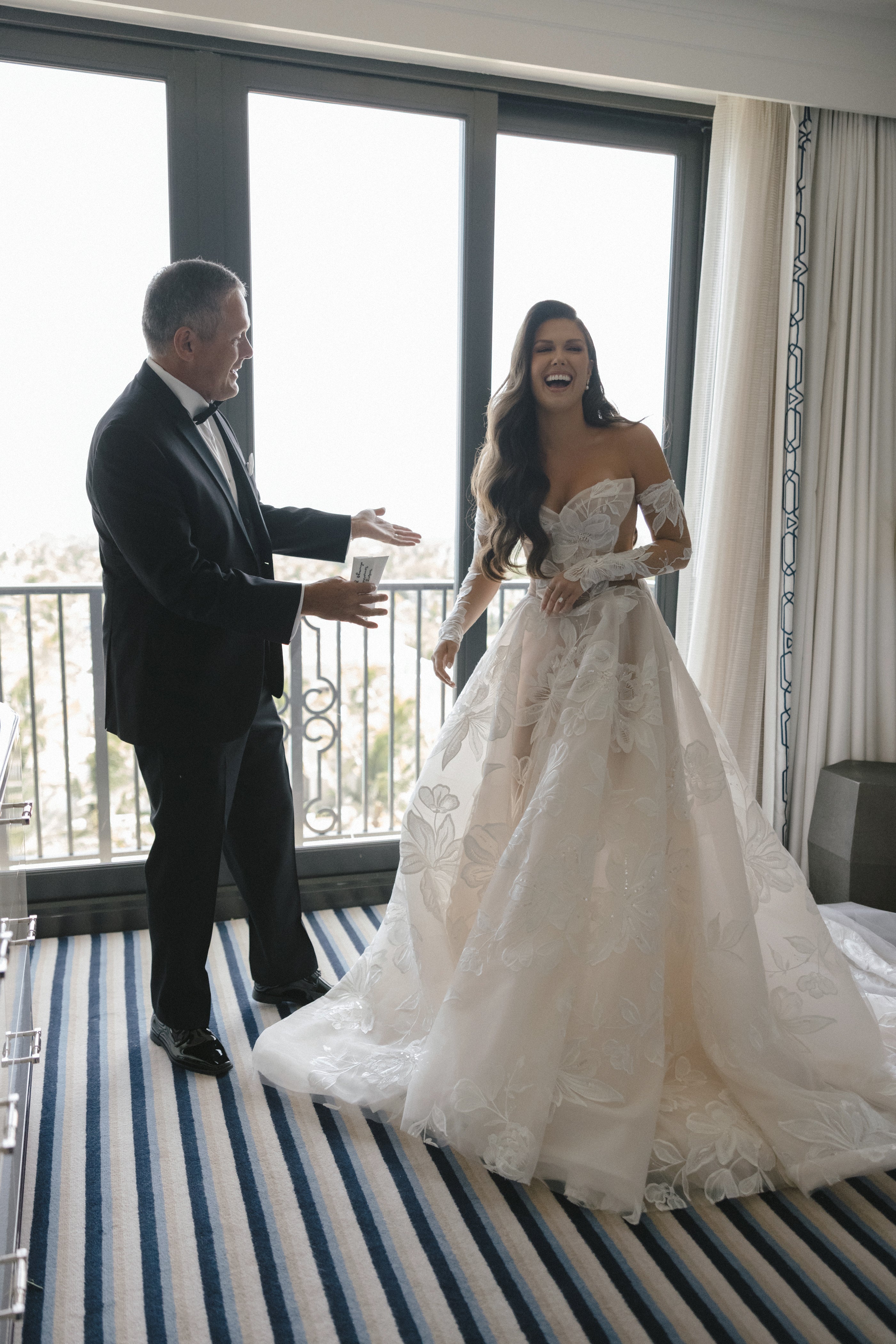 Man and woman in formal attire standing on a balcony with large windows.
