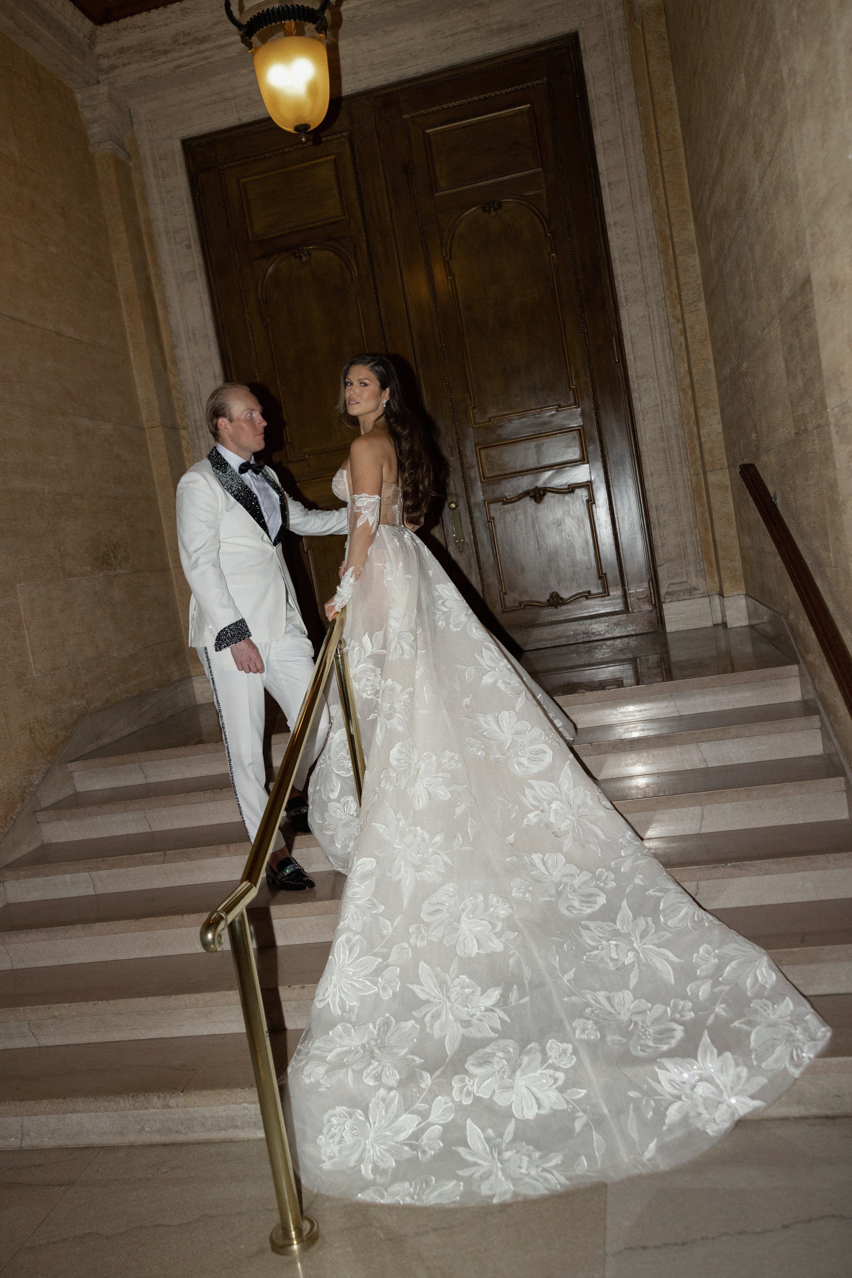 Man and woman in formal attire standing on a staircase with a decorative light fixture above.
